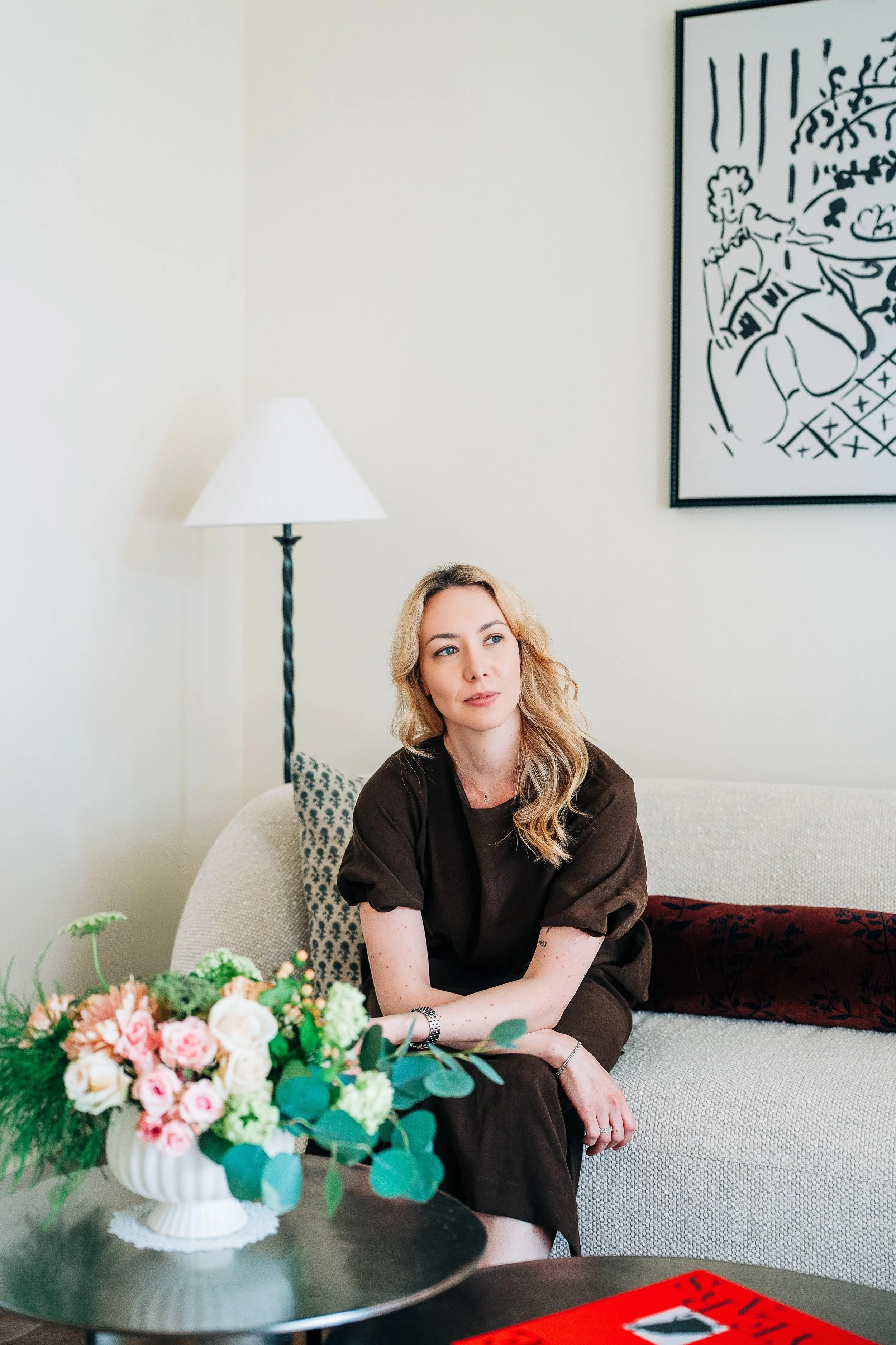 A woman with blonde hair sitting on a light-colored sofa, wearing a dark brown dress, with a colorful bouquet of flowers on a black coffee table in front of her. A large abstract black and white artwork hangs on the wall behind her, with a white table lamp to her left.