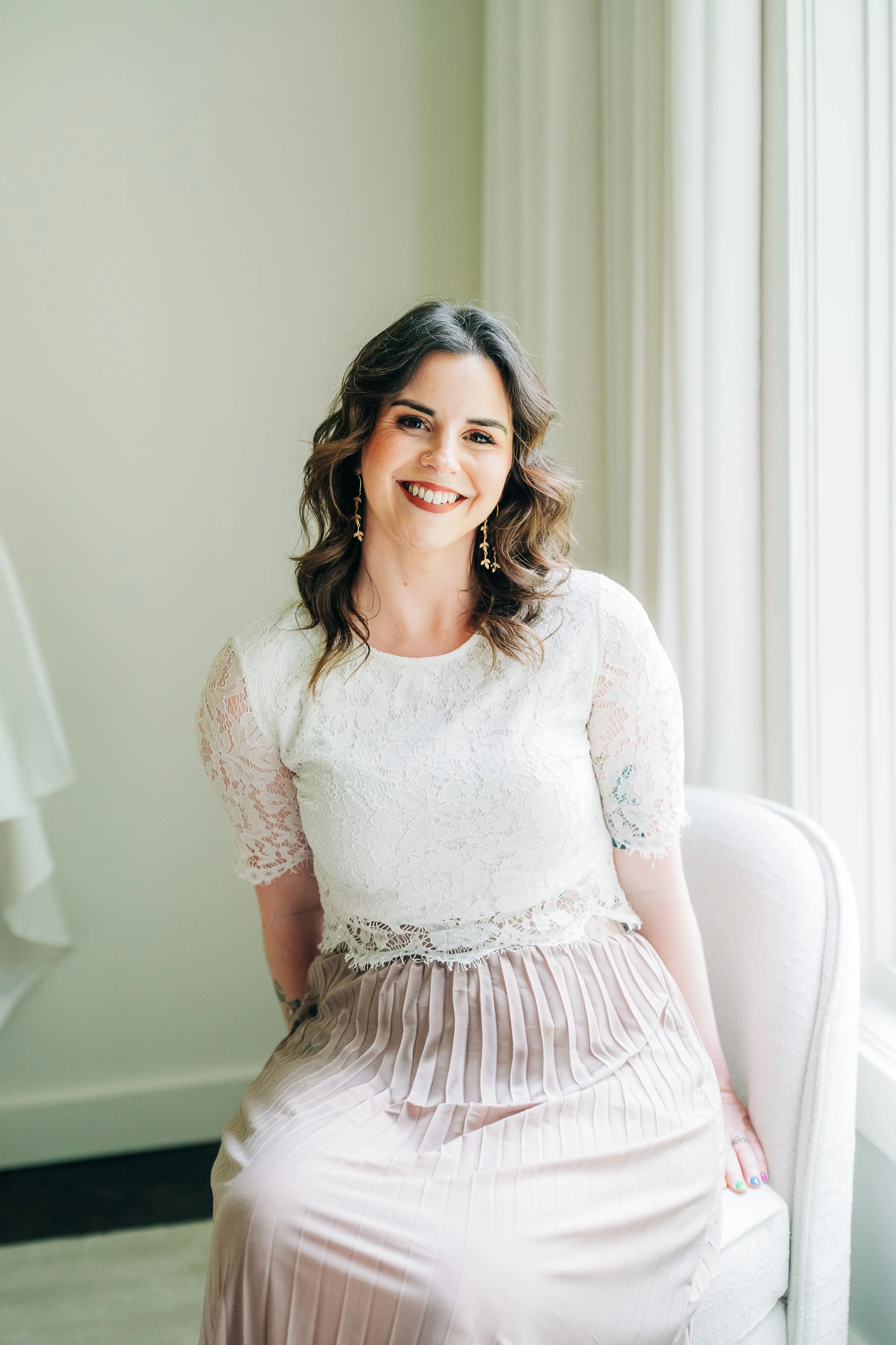 Smiling woman with dark wavy hair, wearing earrings, dressed in a white lace top and a pleated beige skirt, sitting on a cream-colored chair near a window with white curtains, in a softly lit room.