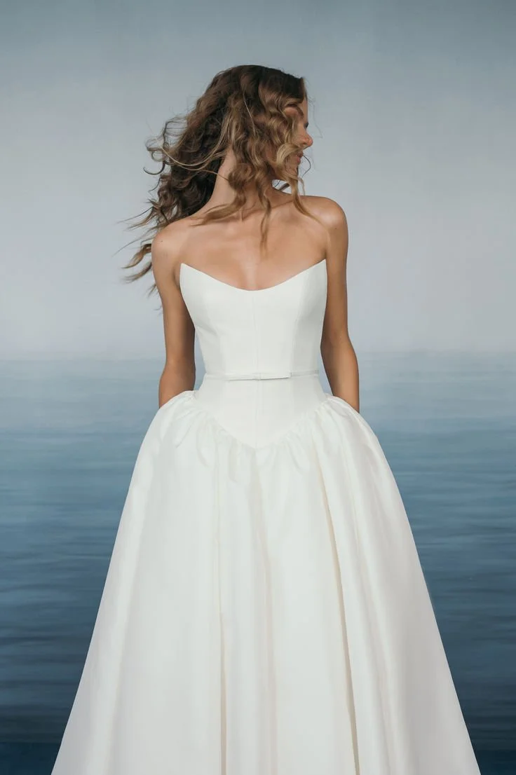 A woman in a white strapless wedding dress standing by the ocean, with her head turned to the side and hair flowing.