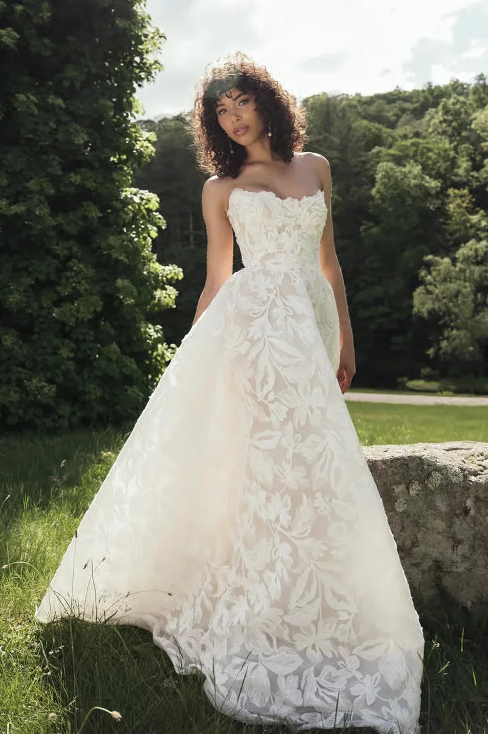 A woman in a strapless white wedding dress standing outdoors in a grassy area with trees in the background.