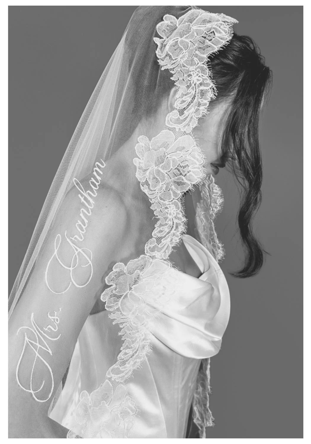 A woman in bridal attire wearing a lace veil with the inscription 'Alex, thank you' embroidered on it, with dark wavy hair and a satin dress, in black and white.