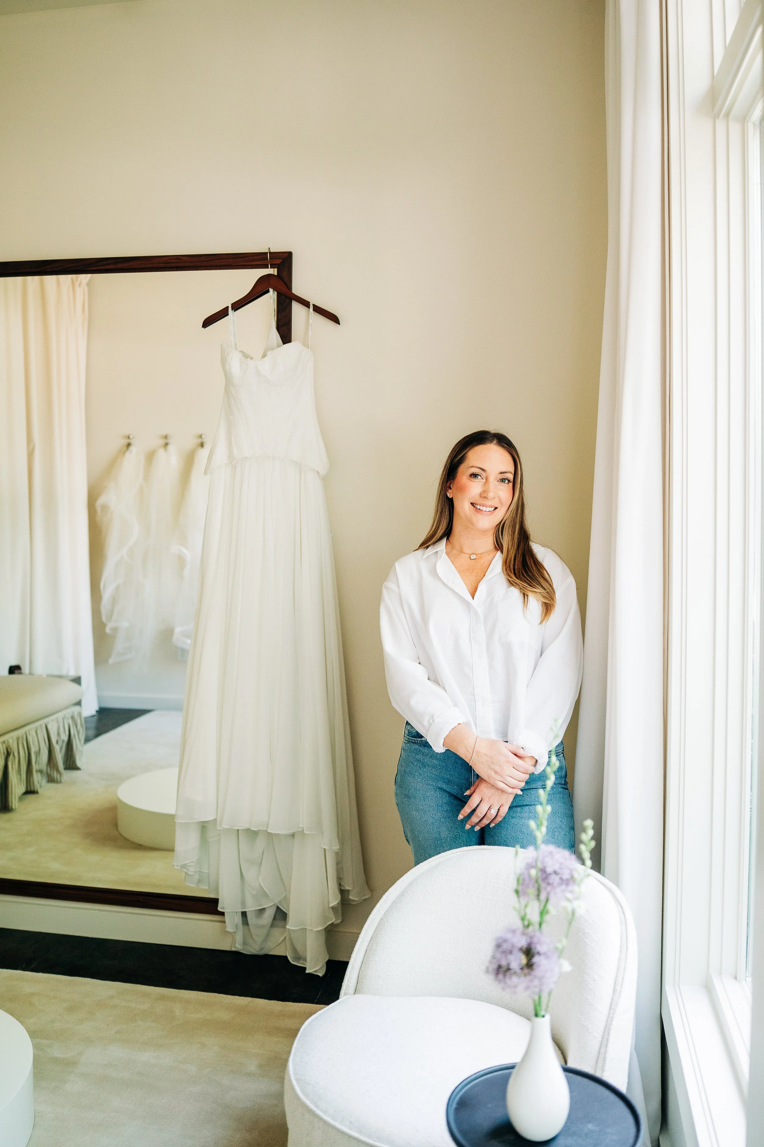 A woman with long hair wearing a white shirt and jeans, standing indoors by a window with white curtains, smiling at the camera. Behind her are two wedding dresses hanging on a mirror and a cream-colored chair with a floral arrangement on a table.