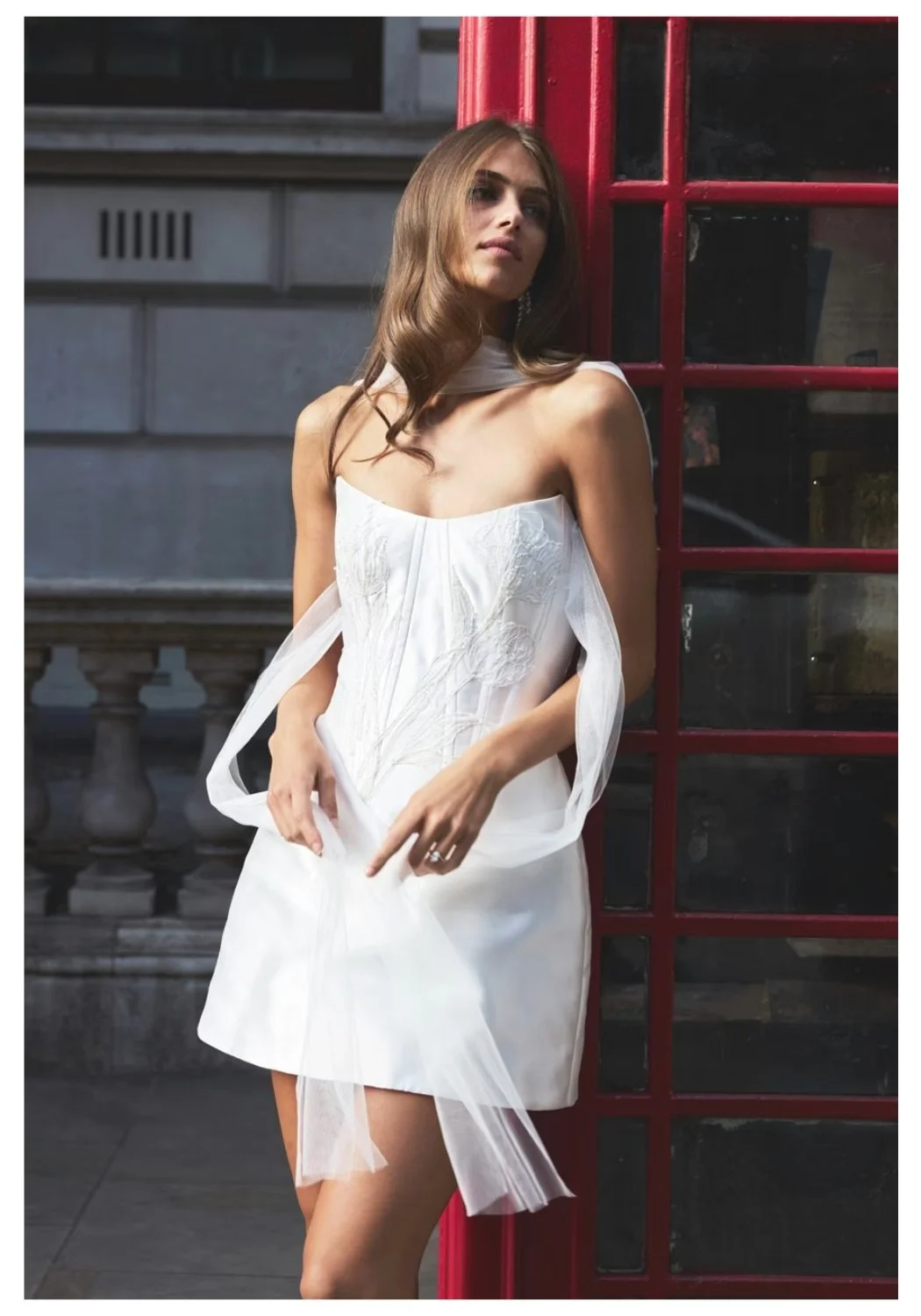 A young woman with long brown hair wearing a white off-shoulder dress standing next to a red phone booth on a city street.
