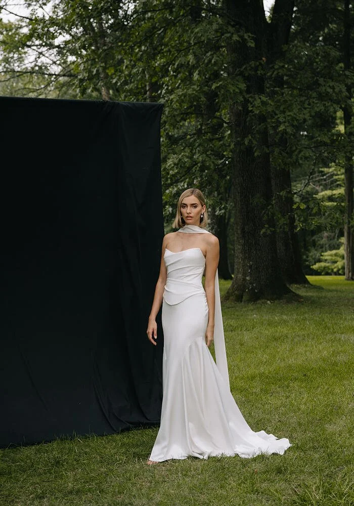 A woman in a strapless white dress standing outdoors near a black backdrop in a lush green park.