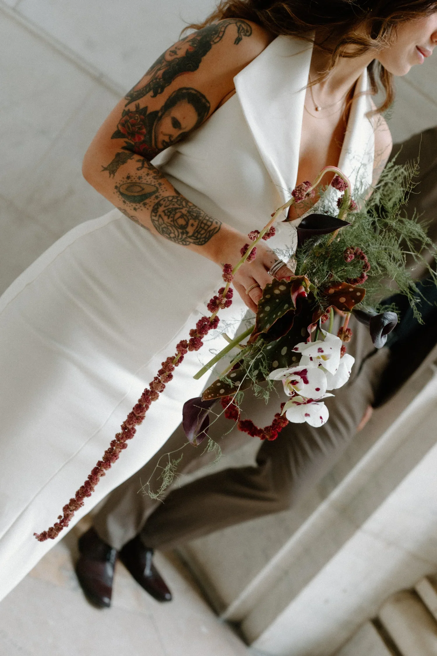 A woman with tattoos on her arm is holding a bouquet of assorted flowers, including orchids and foliage, while wearing a white dress.
