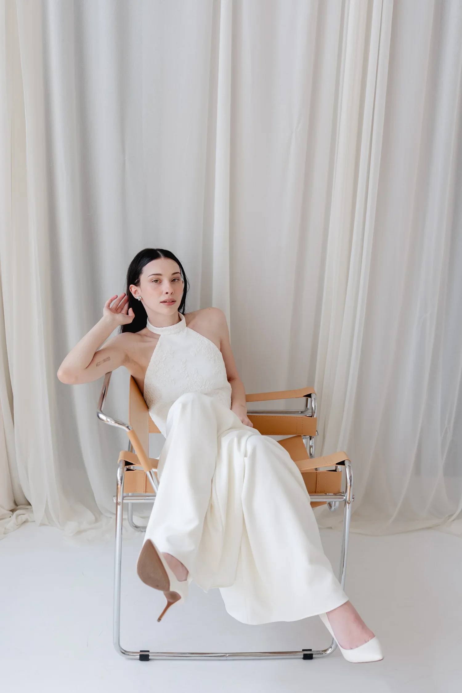 A woman sitting on a modern chair in front of cream curtains. She is wearing a white dress and white heels, with dark hair and minimal makeup, posing with one hand near her ear and the other resting on her lap.