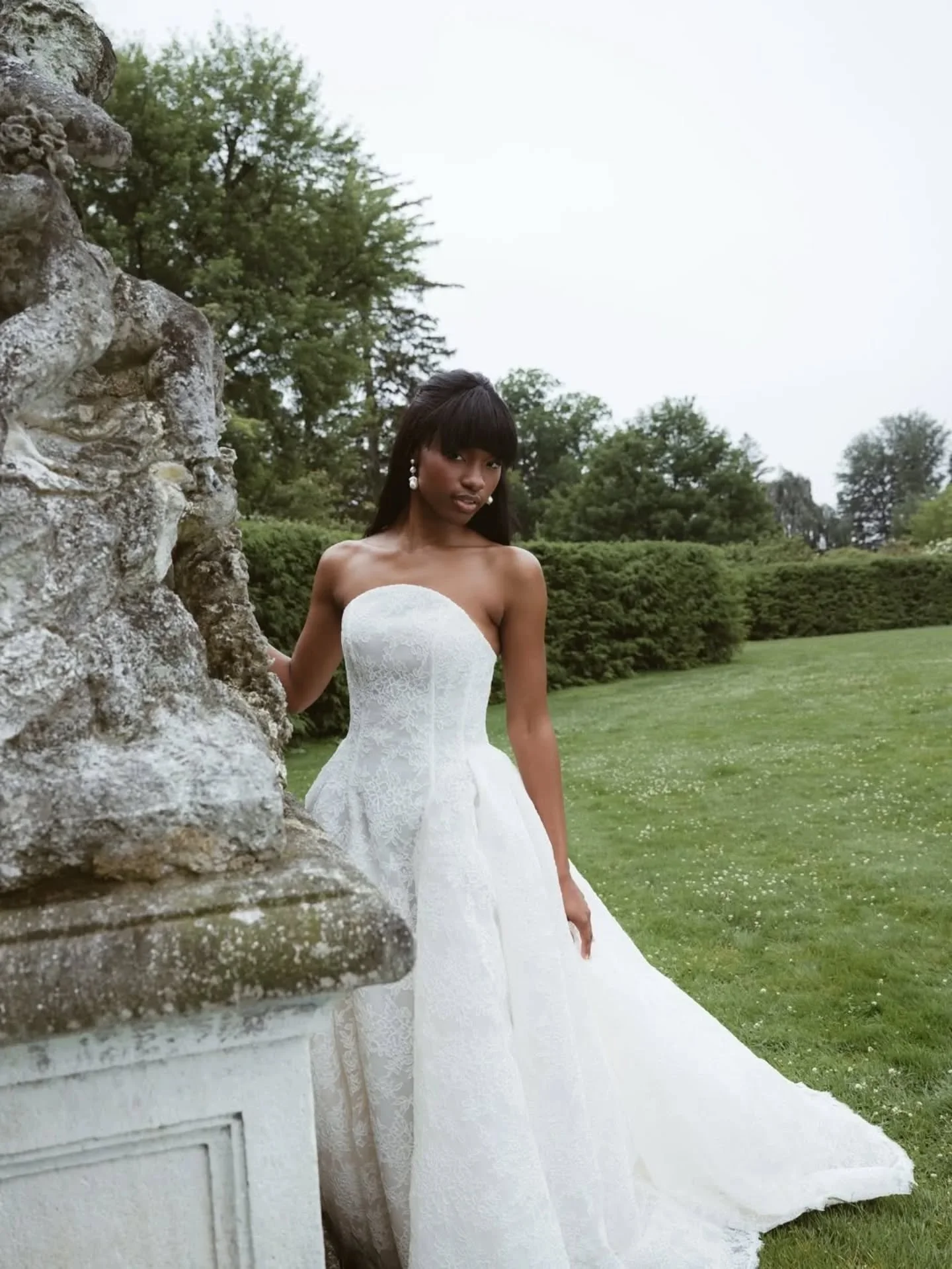 A woman in a white strapless wedding dress with lace details, standing outdoors beside a stone wall with greenery and trees in the background.