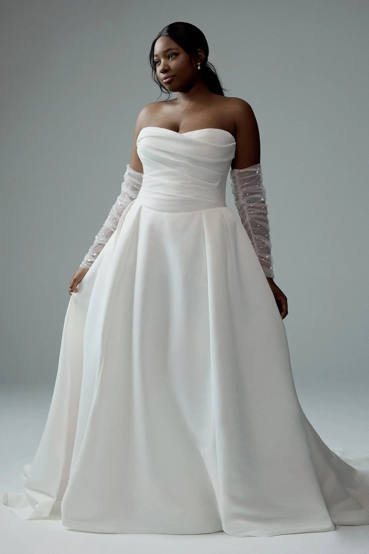 A woman wearing an elegant strapless white wedding gown with a full skirt and sheer, ruched, beaded long sleeves, standing against a plain, light grey background.