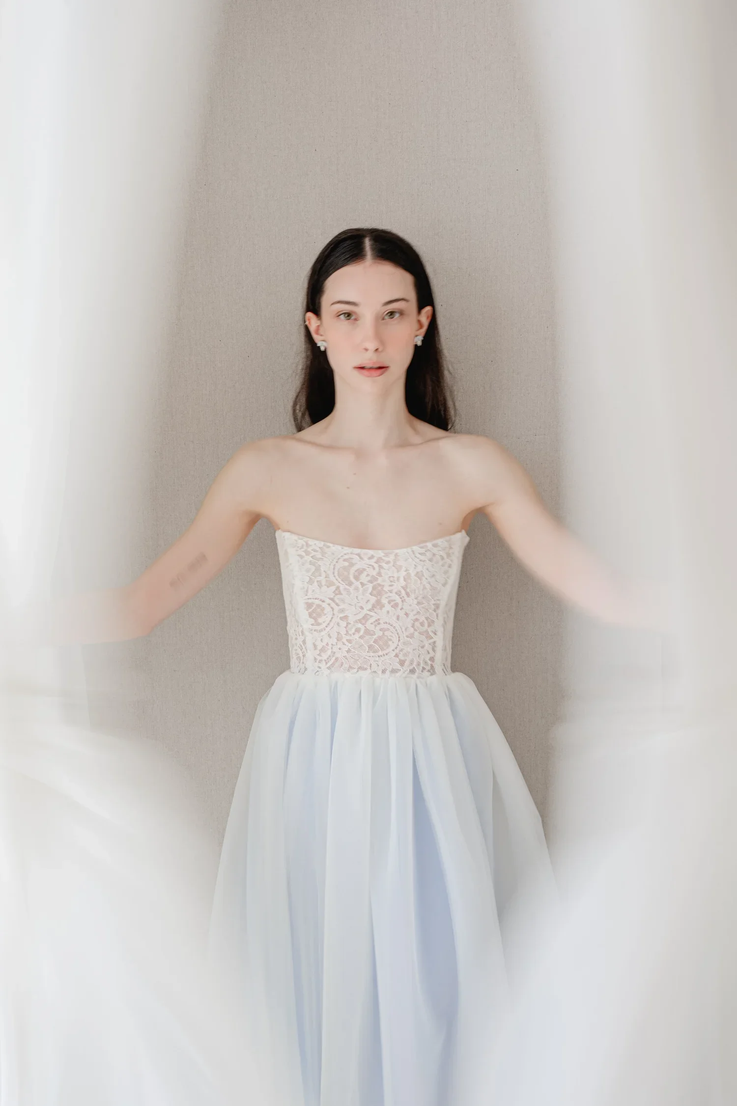 A young woman with dark hair and earrings, wearing a strapless white lace dress with a voluminous skirt, standing against a plain beige wall.