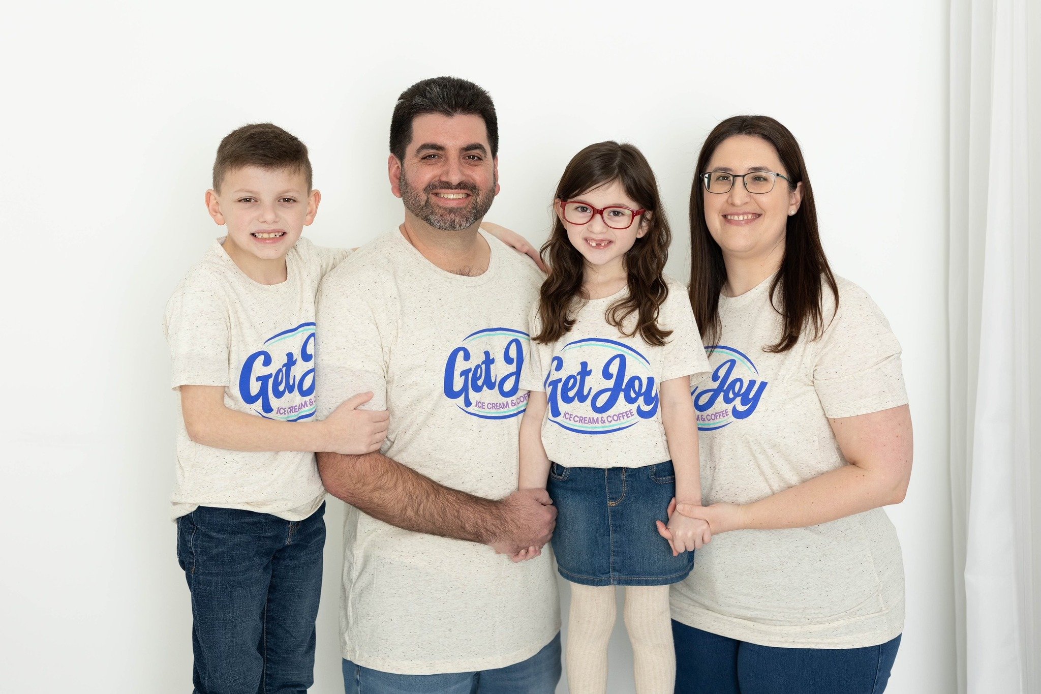 Family of four smiling and wearing matching T-shirts that say 'Get Joy,' standing against a white background.