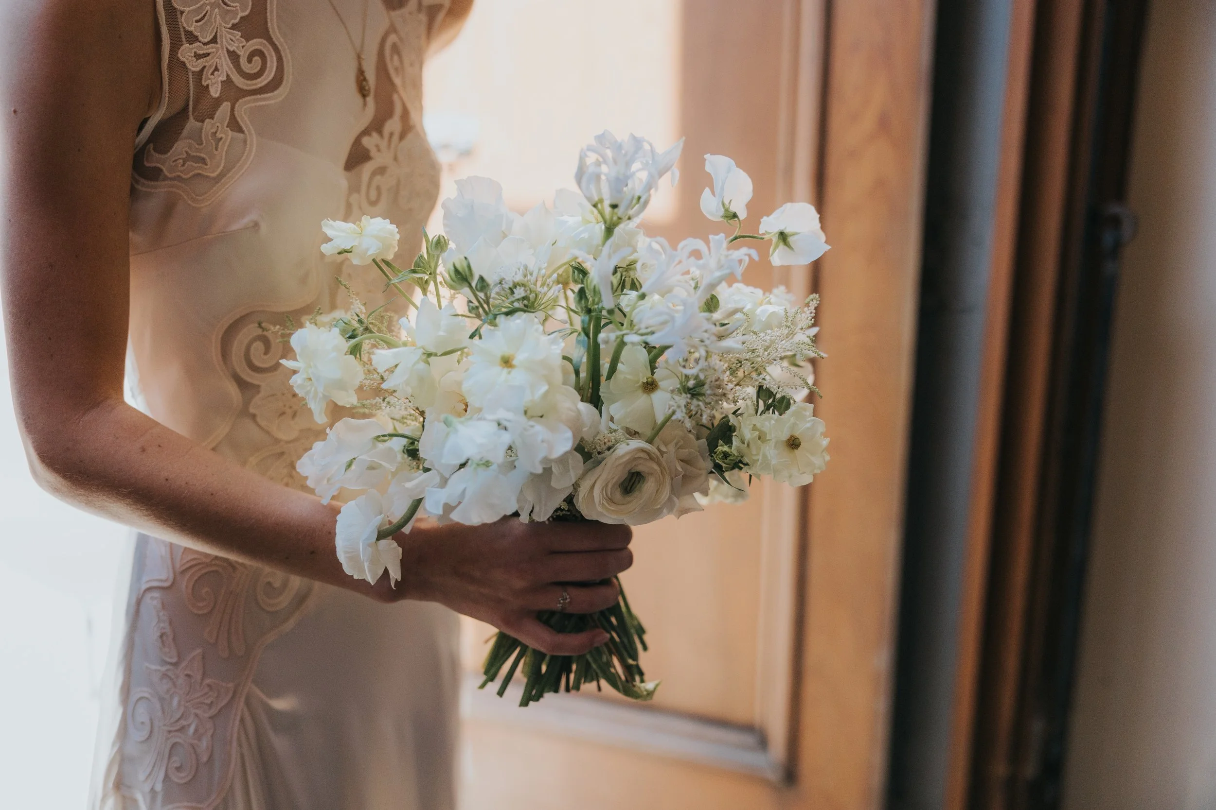 A bride in a detailed lace wedding dress holding a bouquet of white flowers.