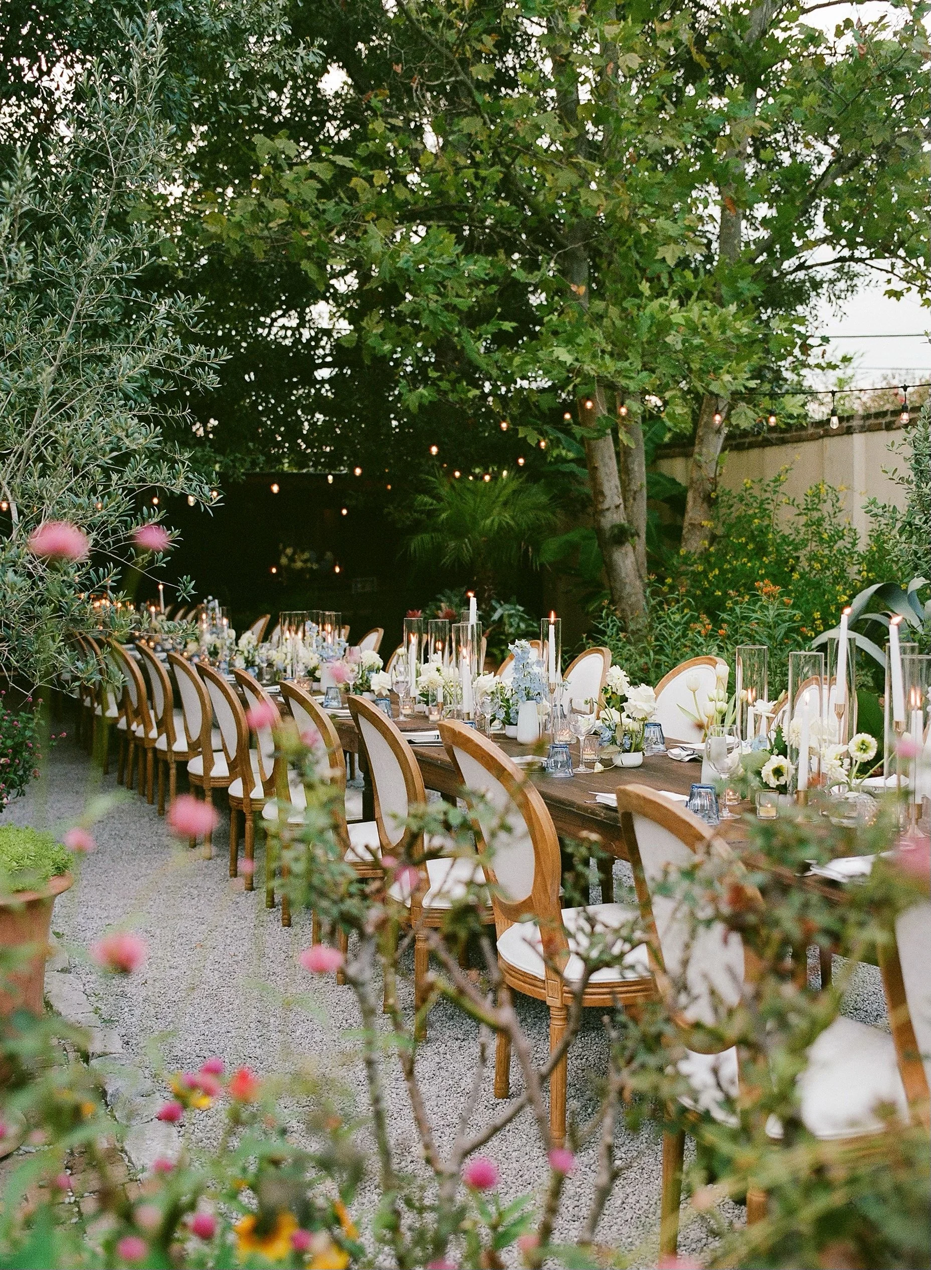 Long tablescape with candlelight and lush floral centerpieces in a tree covered courtyard for an outdoor wedding reception.