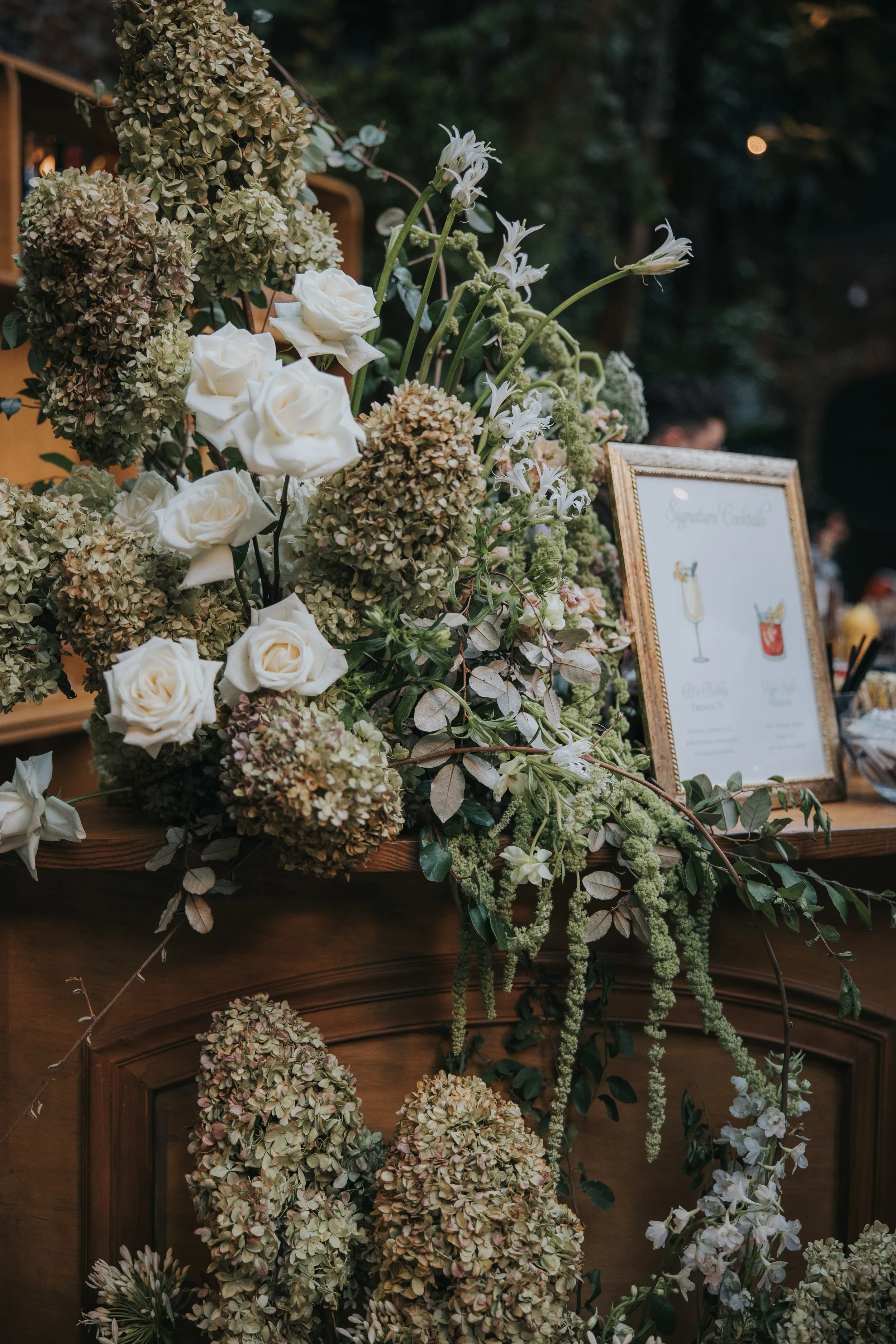Lush bar floral arrangement with hydrangeas, roses, and greenery accenting a custom cocktail menu at a wedding reception.
