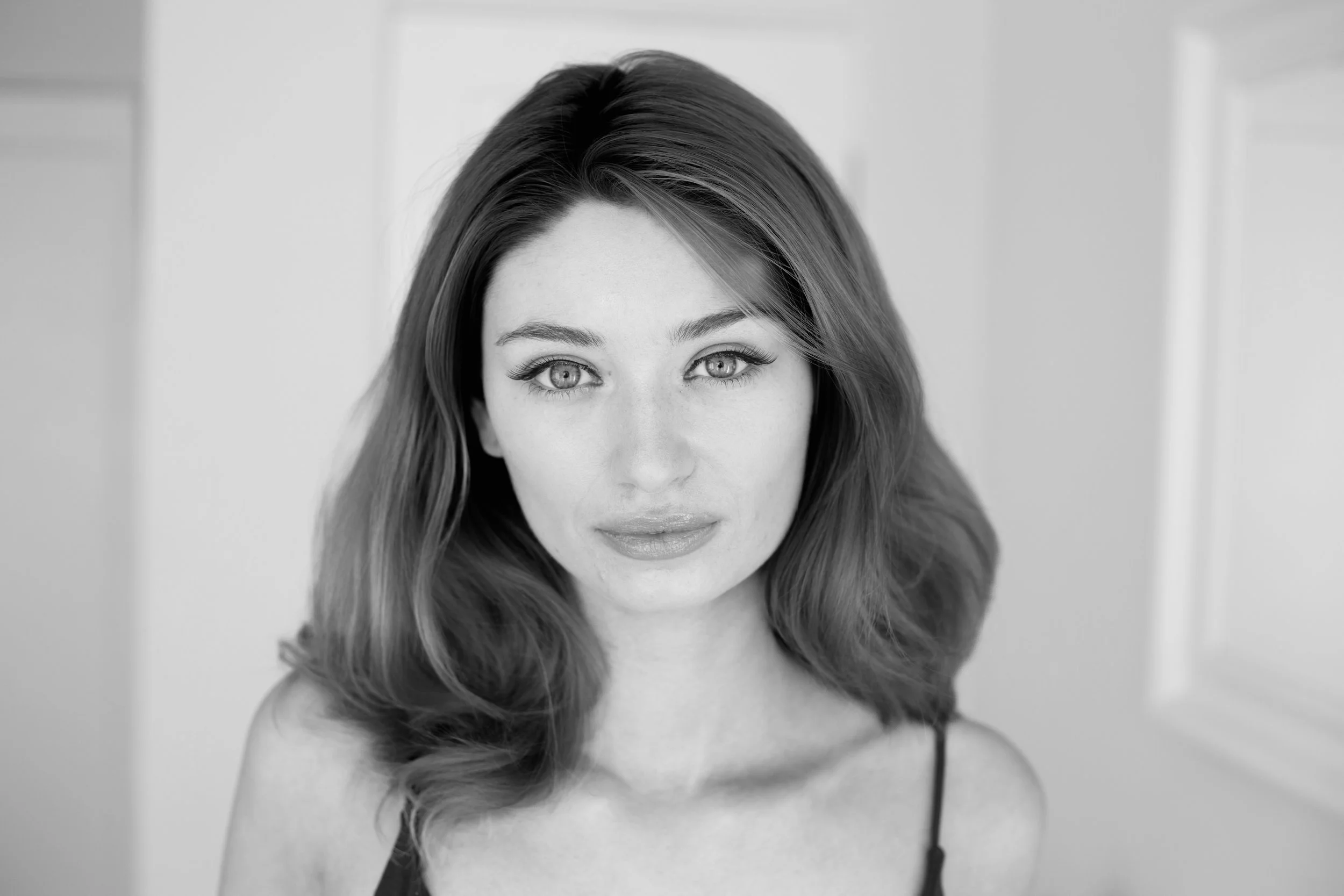 Black and white portrait of an actor, writer, filmmaker, short film director, film producer woman with wavy hair, light eyes, and a subtle beautiful smile, in front of a softly blurred background.