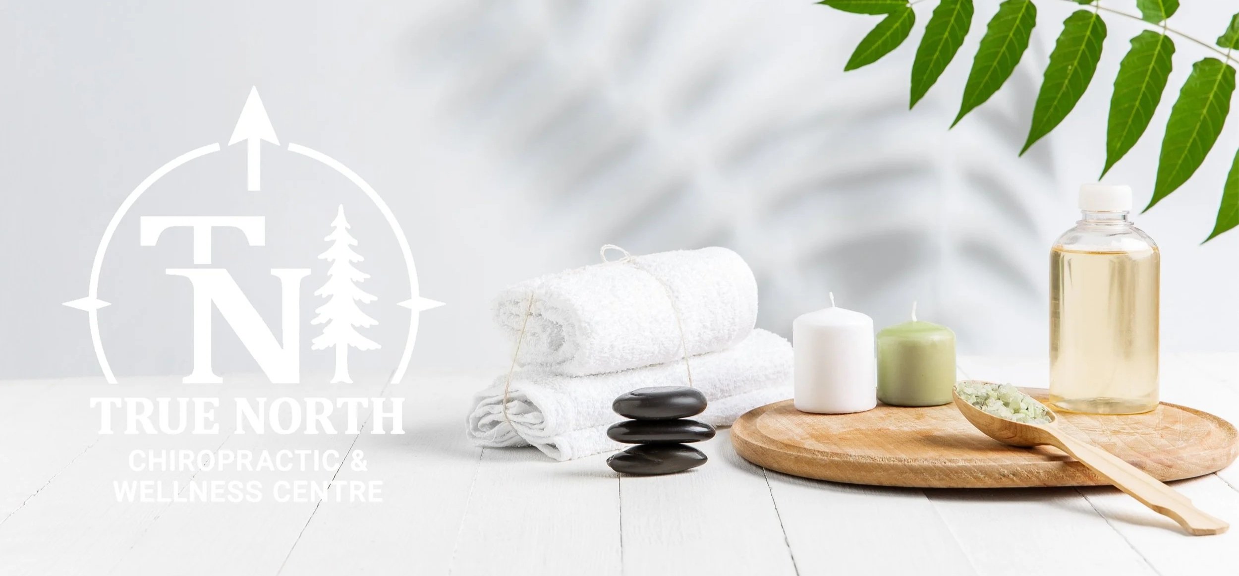 Spa and wellness items including rolled white towels, stacked black stones, white and green candles, a wooden dish with herbal mixture, a small bottle of oil, and green leaves, arranged on a wooden tray.