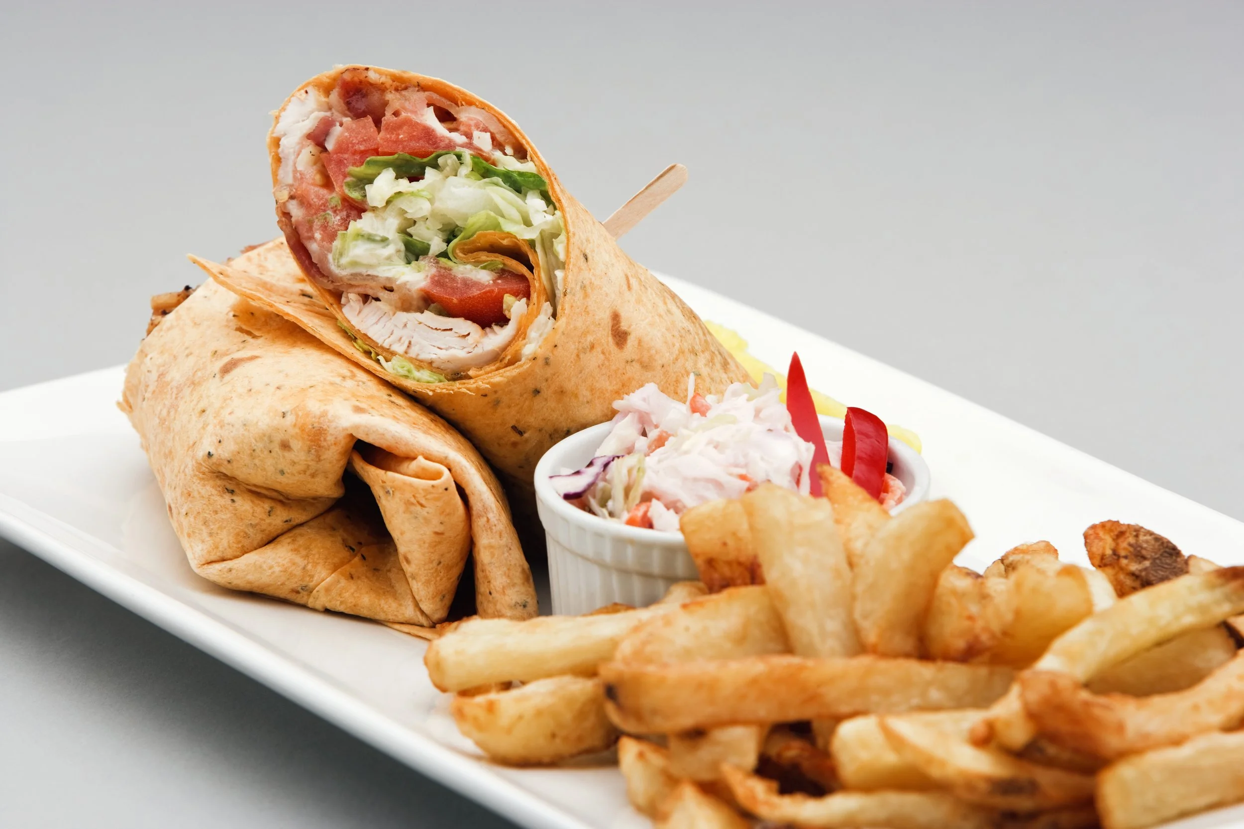 A plate with a burrito, French fries, and coleslaw, with the burrito partially unwrapped to show lettuce, tomato, and chicken inside.