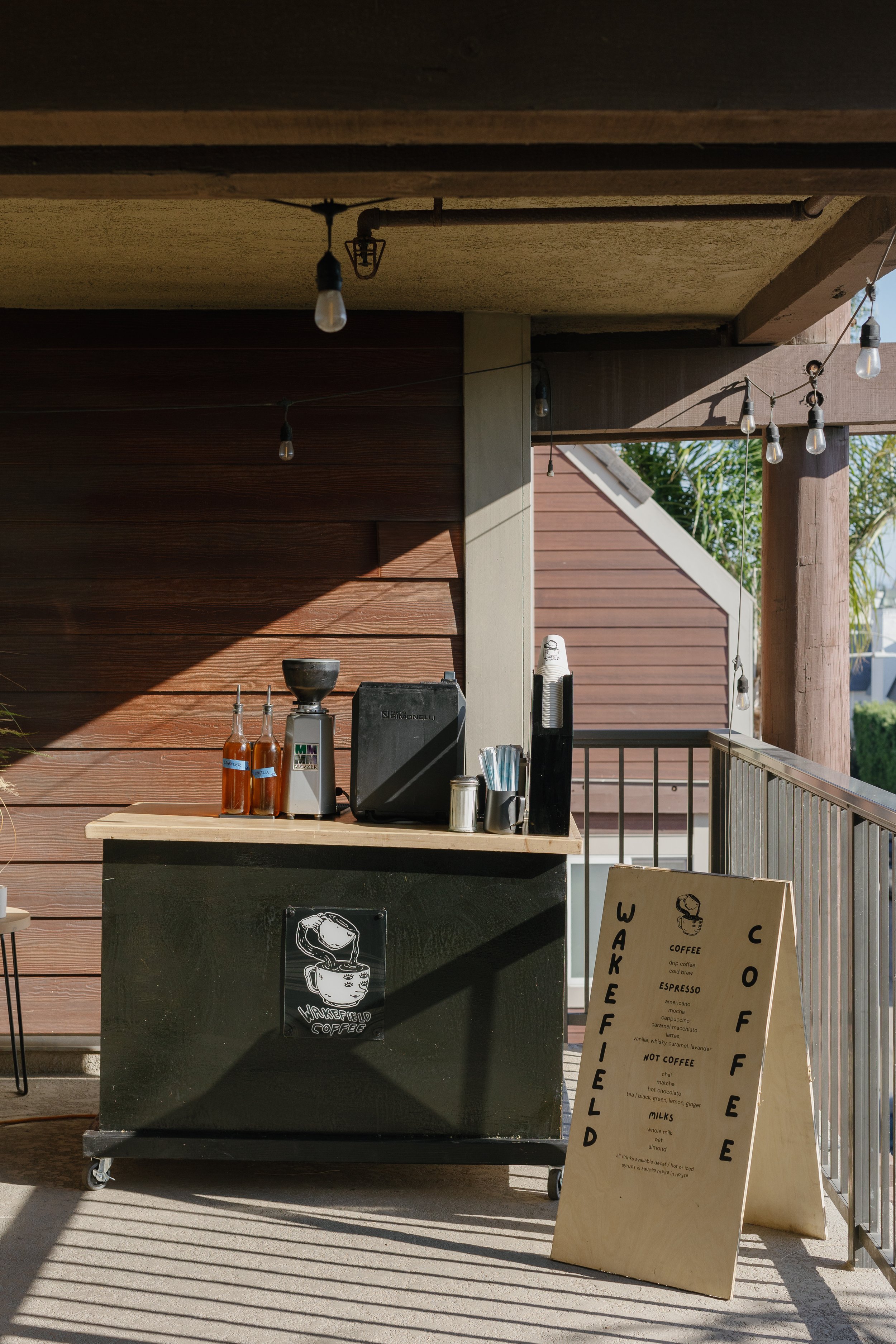 Coffee stand on a balcony with black and wooden elements, sign listing coffee and milk options, string lights overhead, and a partial view of a house with wooden siding in the background.