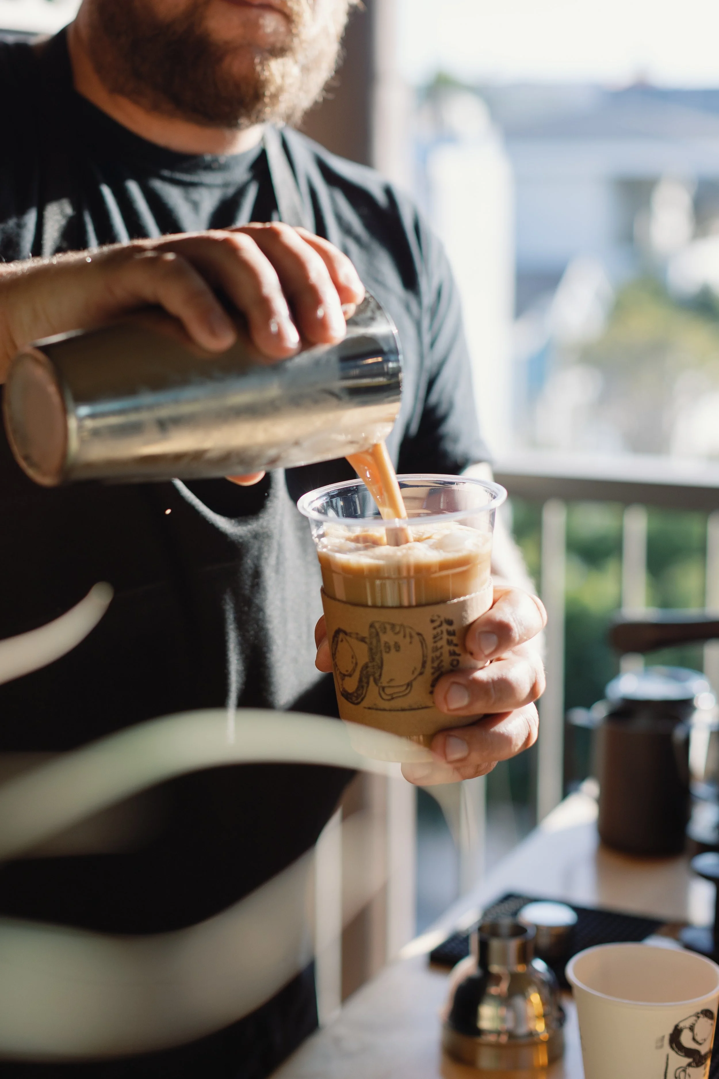 Person pouring coffee from a shaker into a clear plastic cup with a cardboard sleeve, on a sunny day in a cafe.