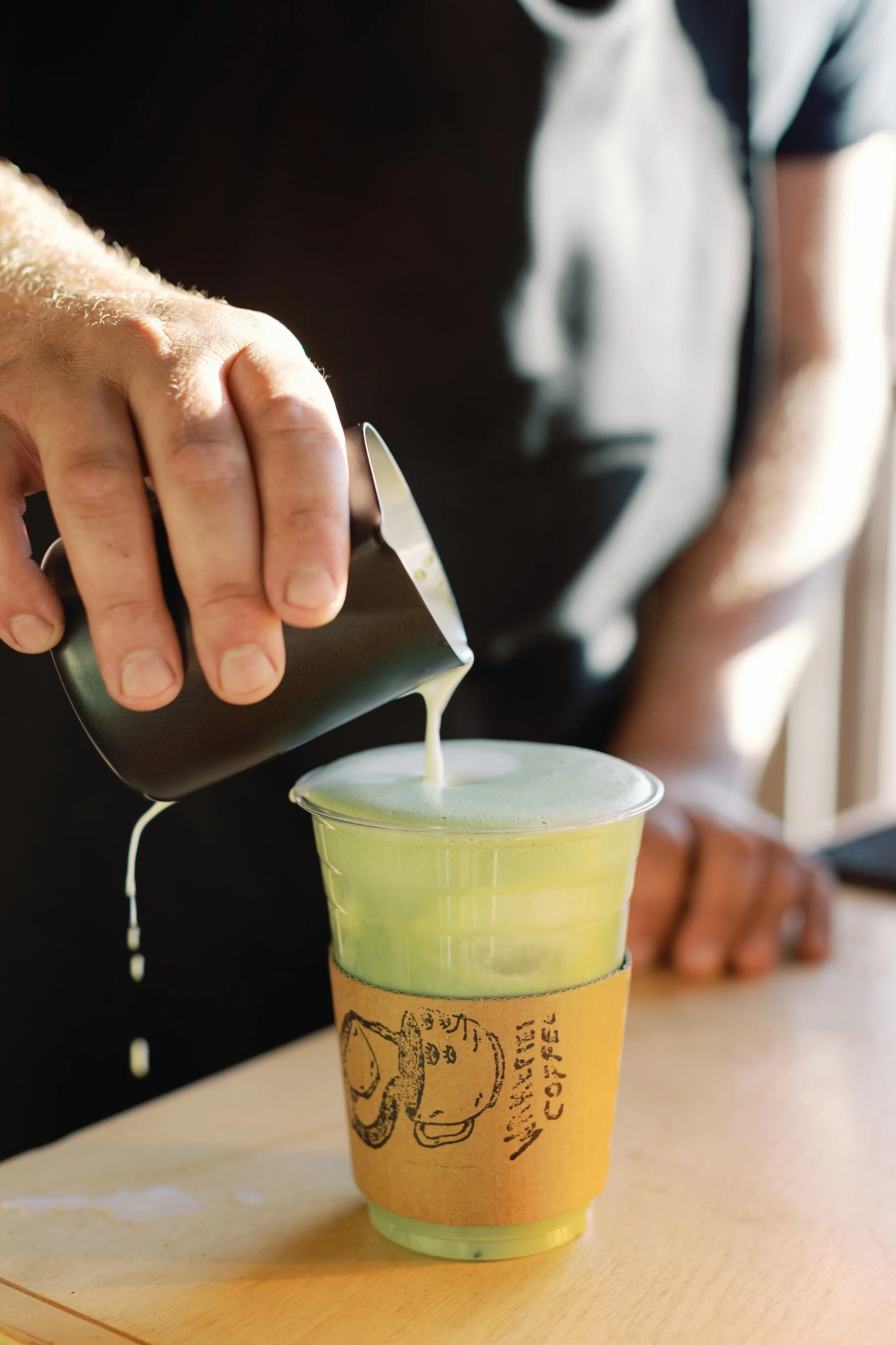 A person pouring a green beverage from a can into a clear plastic cup with a cardboard sleeve that has a coffee logo on it.