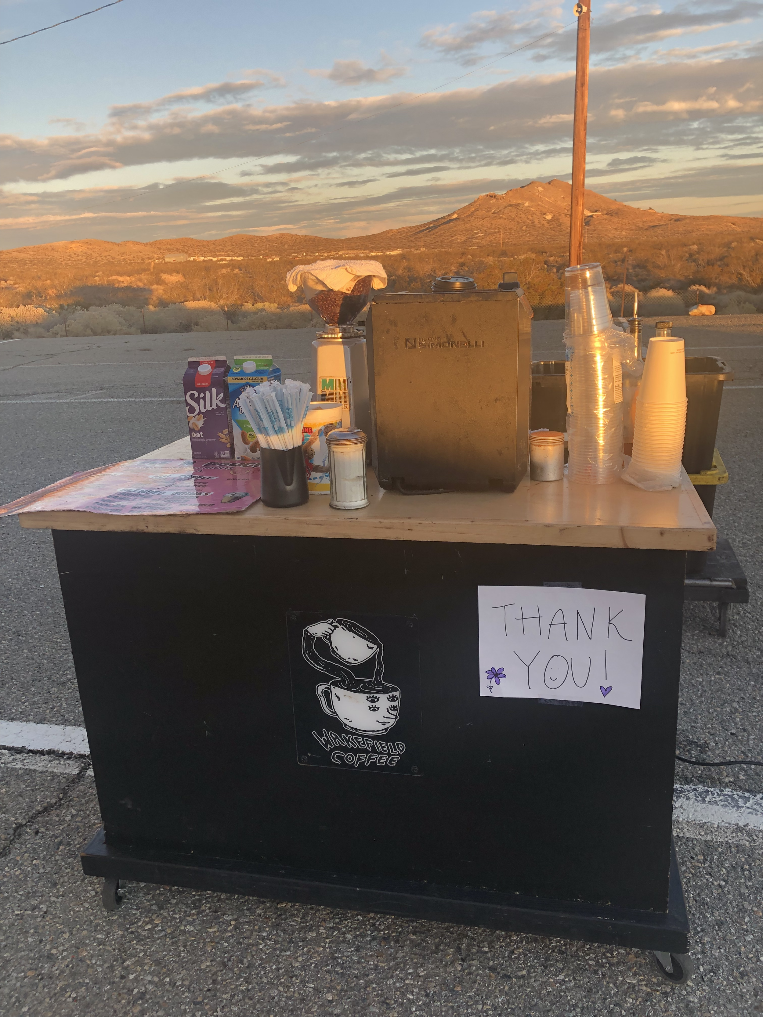 Coffee stand on roadside with mountain and sunset in background, thank you sign and coffee logo on front.