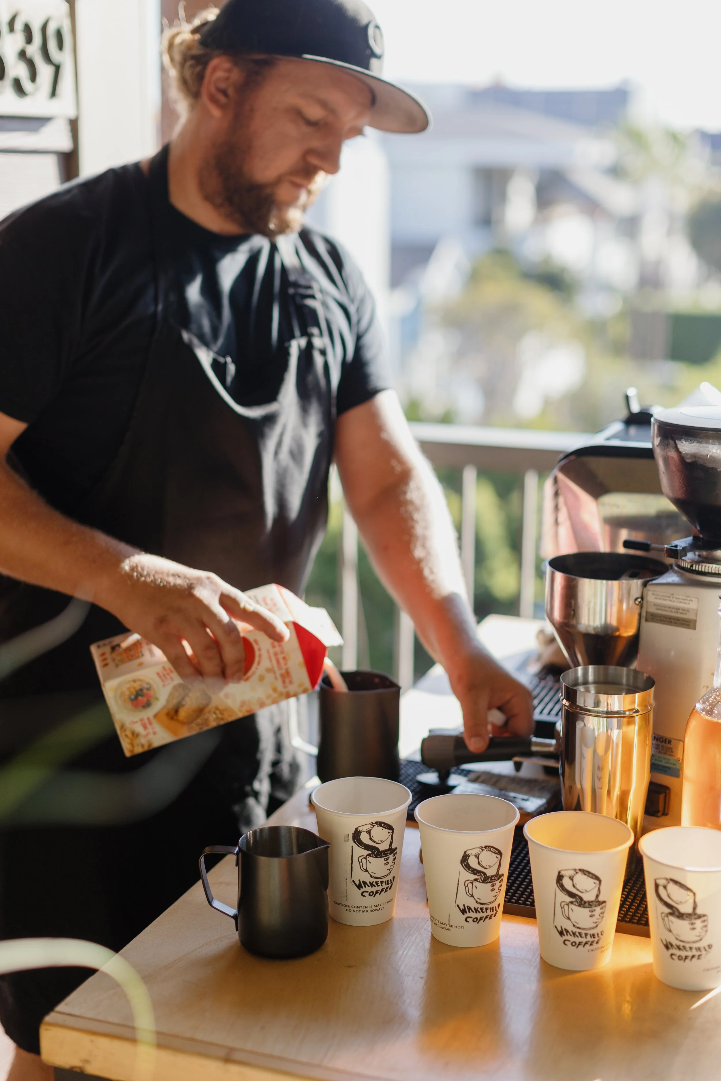Barista preparing coffee behind a counter with disposable cups, coffee equipment, and a bag of coffee mix, on an outdoor patio with sunlight.