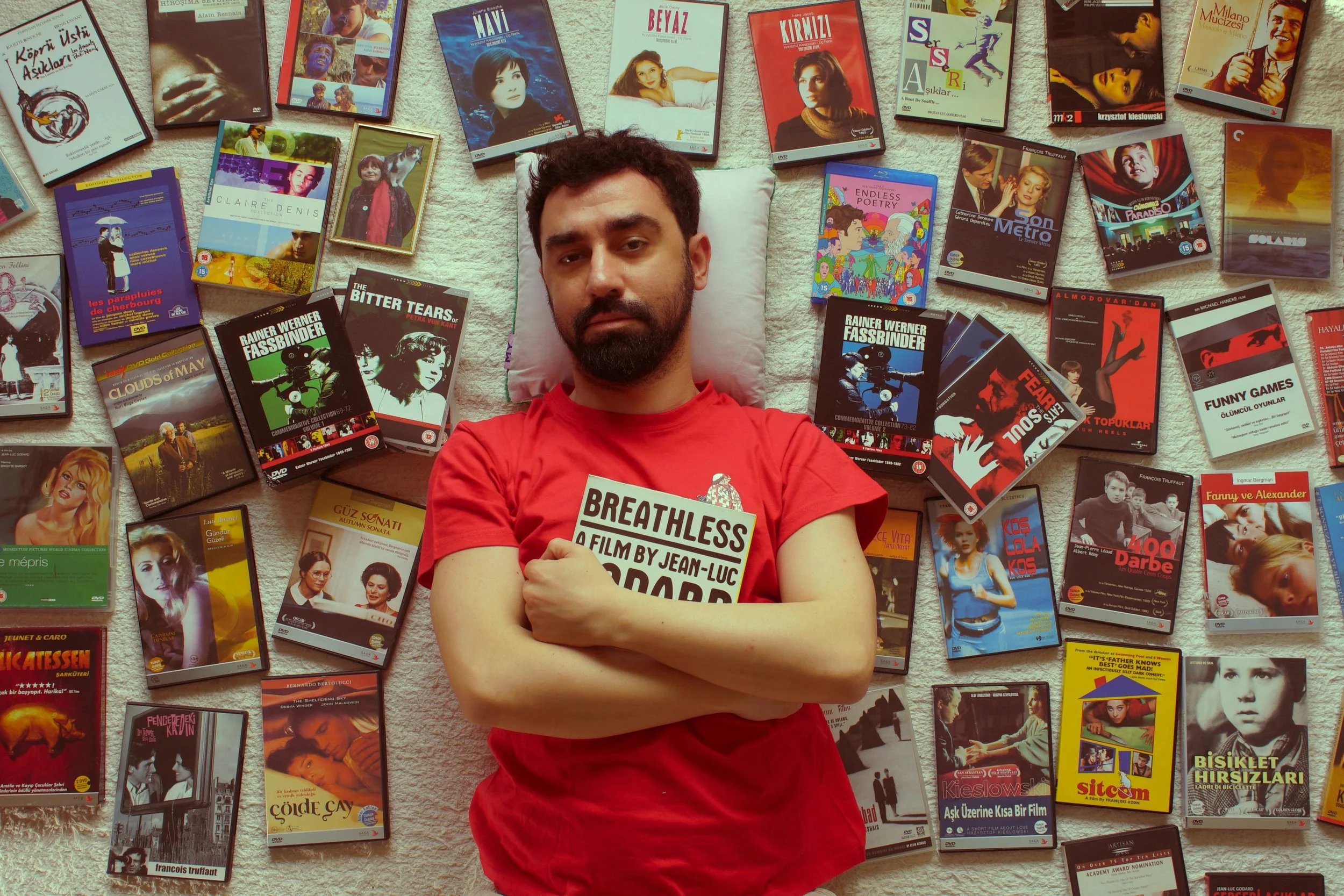 A man with dark hair and a beard lying on a carpeted floor with his arms crossed, holding a newspaper. He is wearing a red T-shirt. Surrounding him are numerous DVDs and books, many with Turkish titles and covers, spread out on the floor.