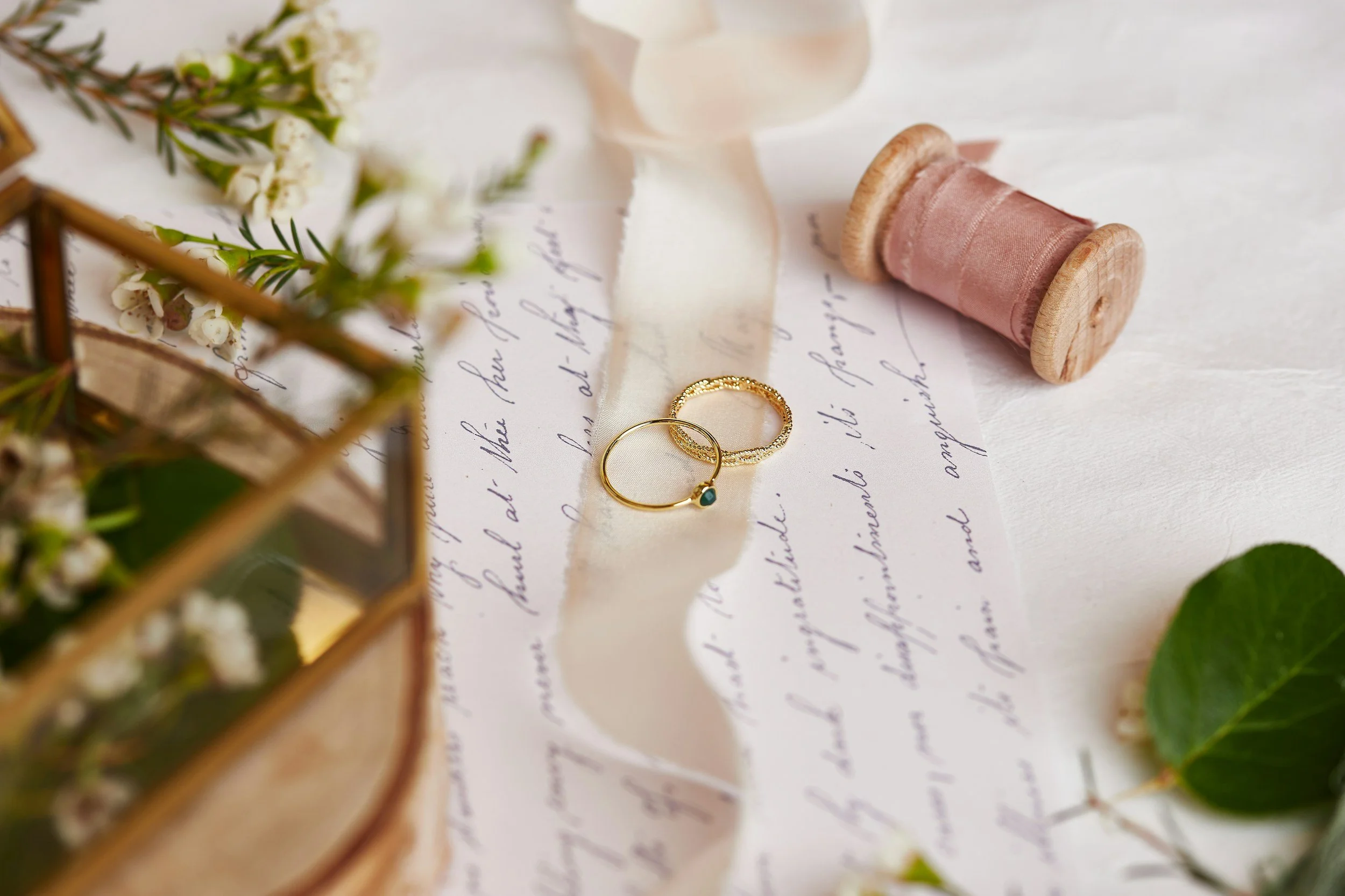 Two gold rings placed on handwritten letter surrounded by flowers, a wooden spool of pink thread, and greenery.