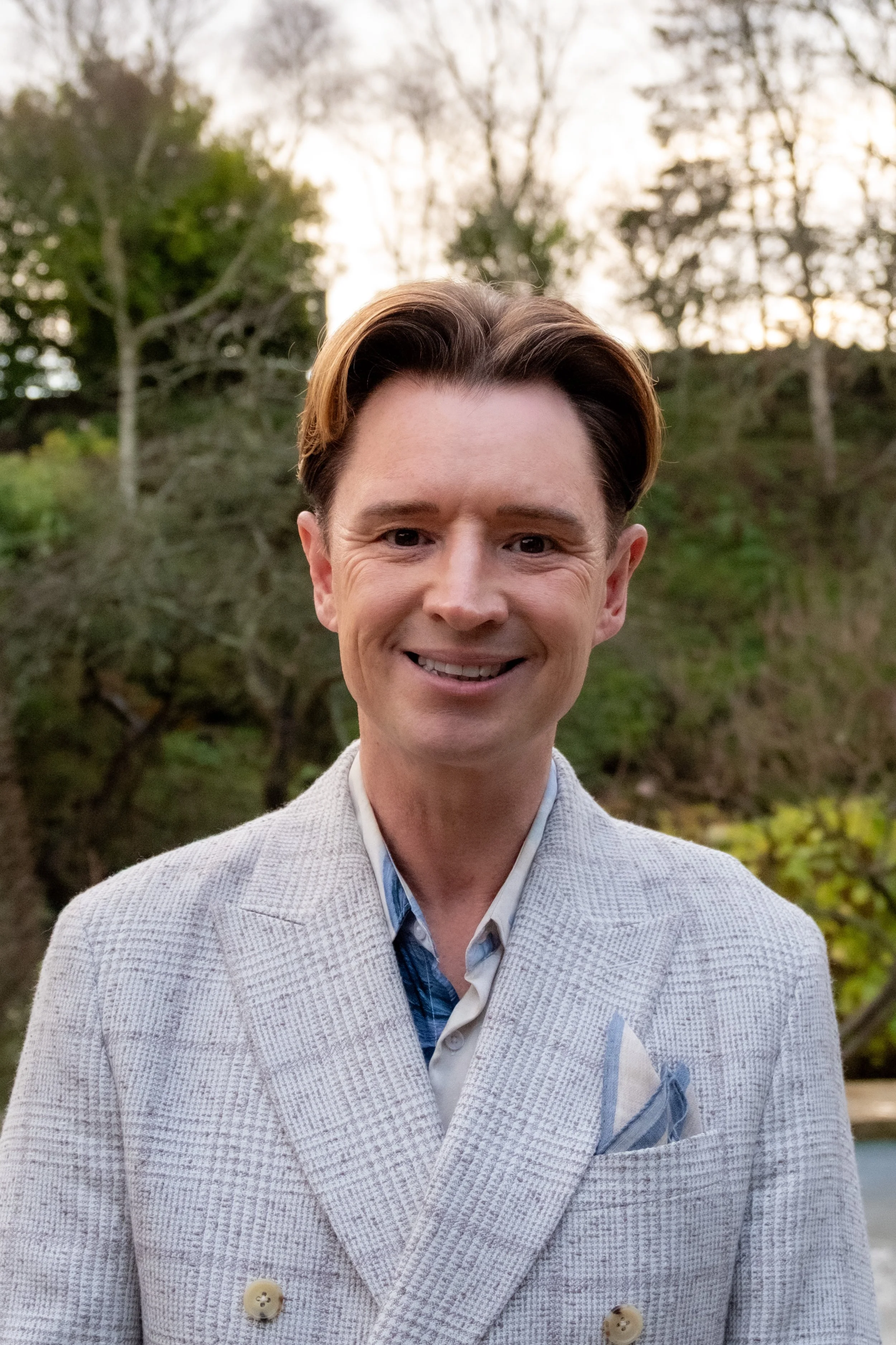 A man in a light-colored checkered blazer and a blue collared shirt standing outdoors in a natural setting with trees and a river in the background.