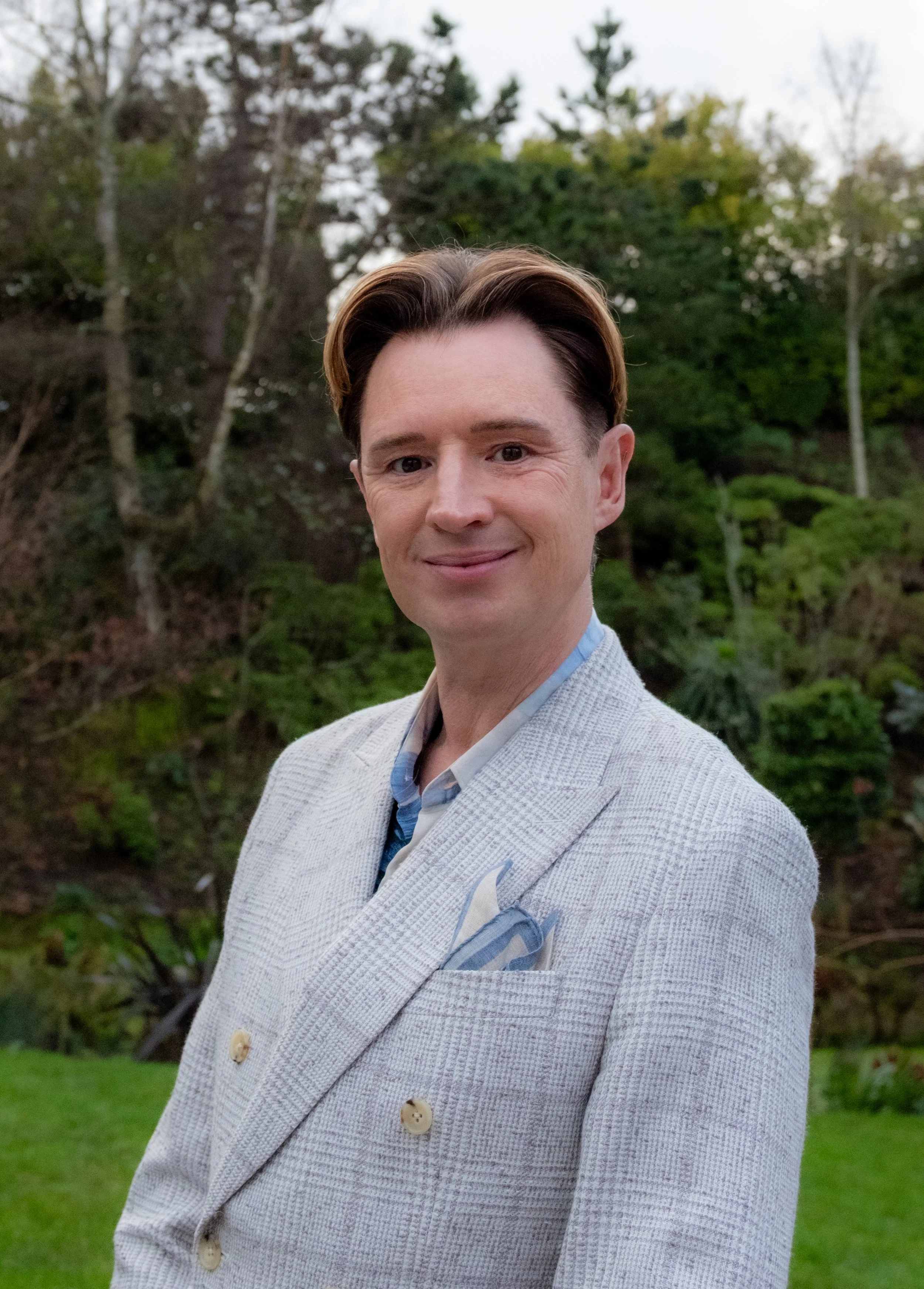 A man with styled brown hair dressed in a light-colored blazer with a pocket square, standing outdoors in front of a backdrop of trees and greenery.