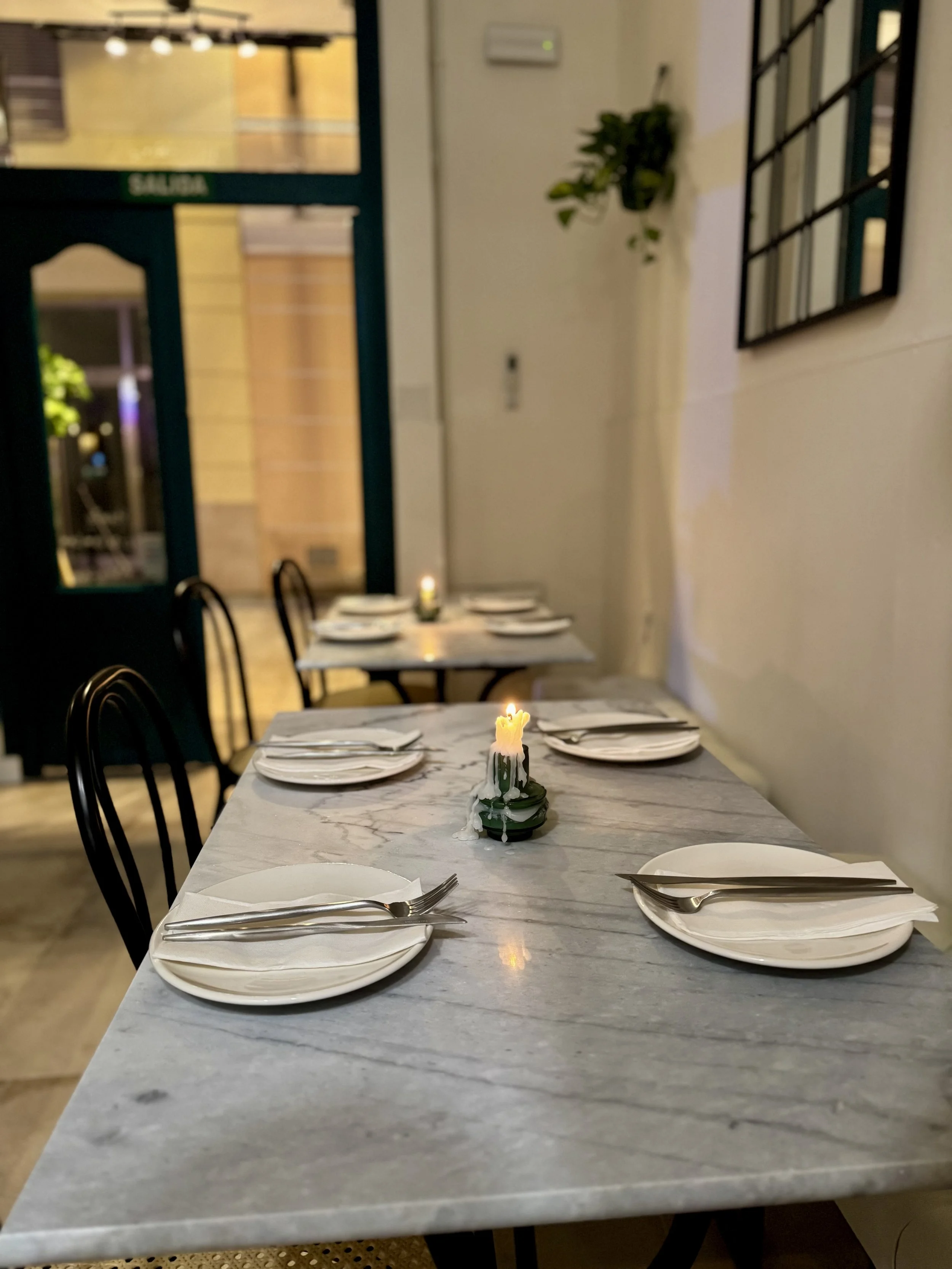 A dining table set for four with white plates, silverware, and a lit candle in the center, inside a cozy restaurant with minimalistic decor and a candlelight ambiance.