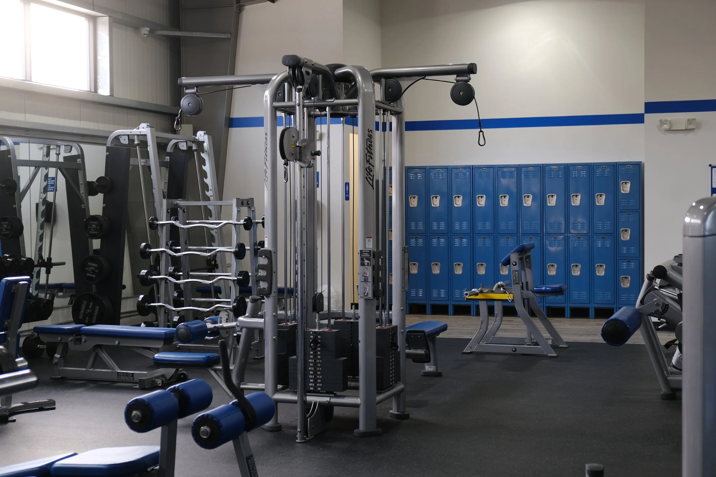 A gym with various workout equipment, including a seated bench, a weight rack with dumbbells, a cable machine, and blue lockers in the background.