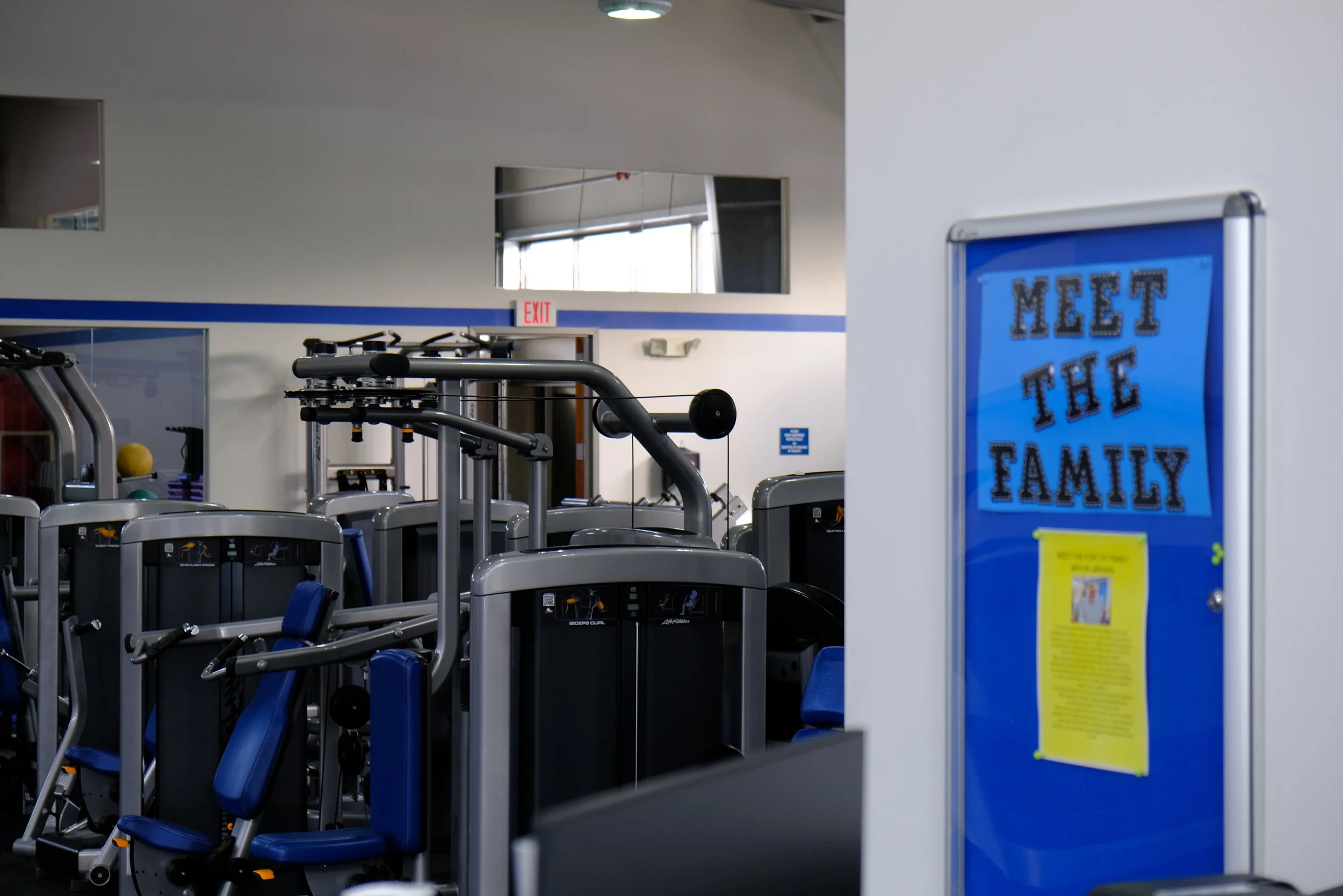 Interior of a gym with various exercise machines, and a blue bulletin board on the wall with a sign saying 'Meet the Family'.