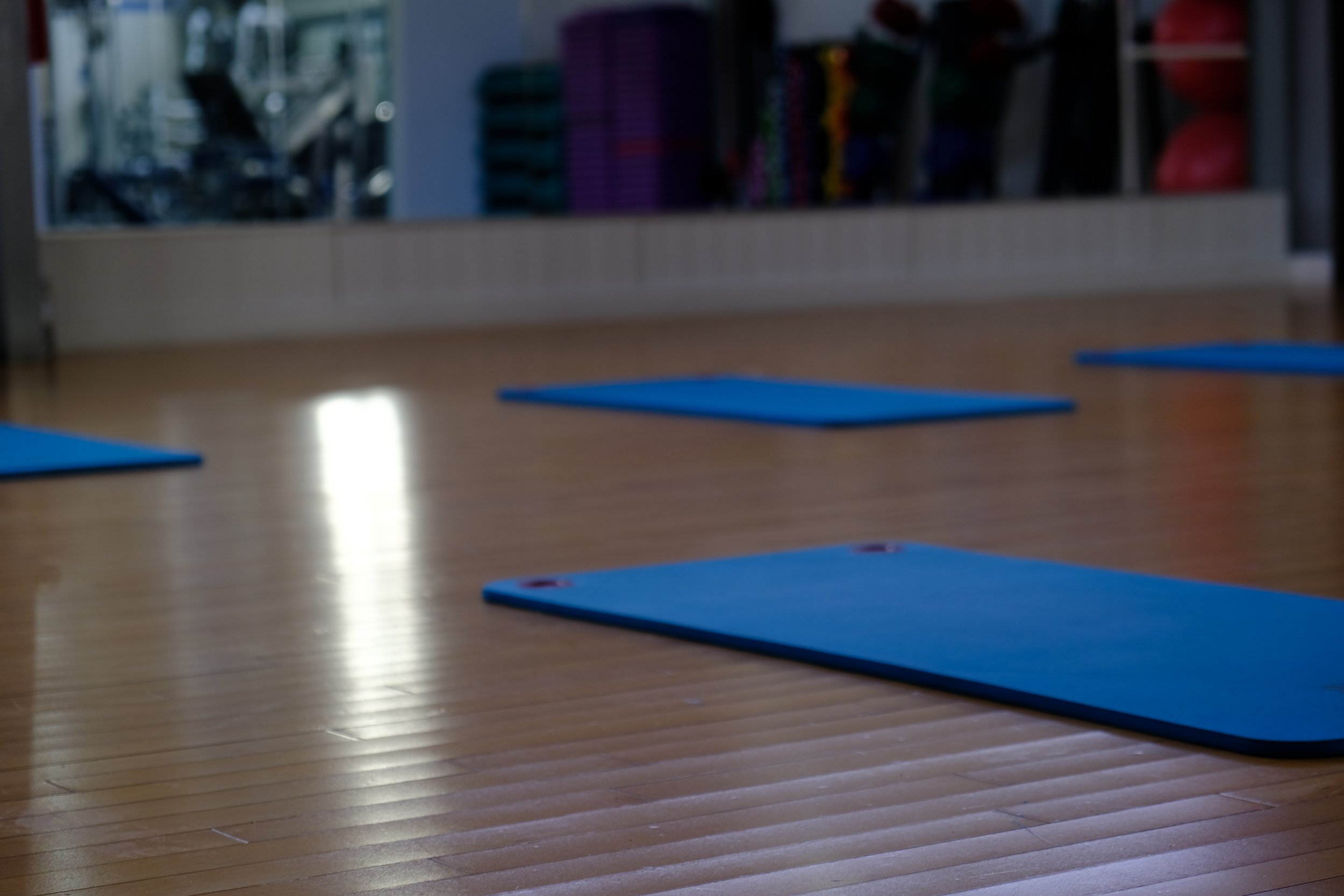 Yoga mats laid out on a wooden floor in a fitness studio near a glass wall with exercise equipment visible in the background.
