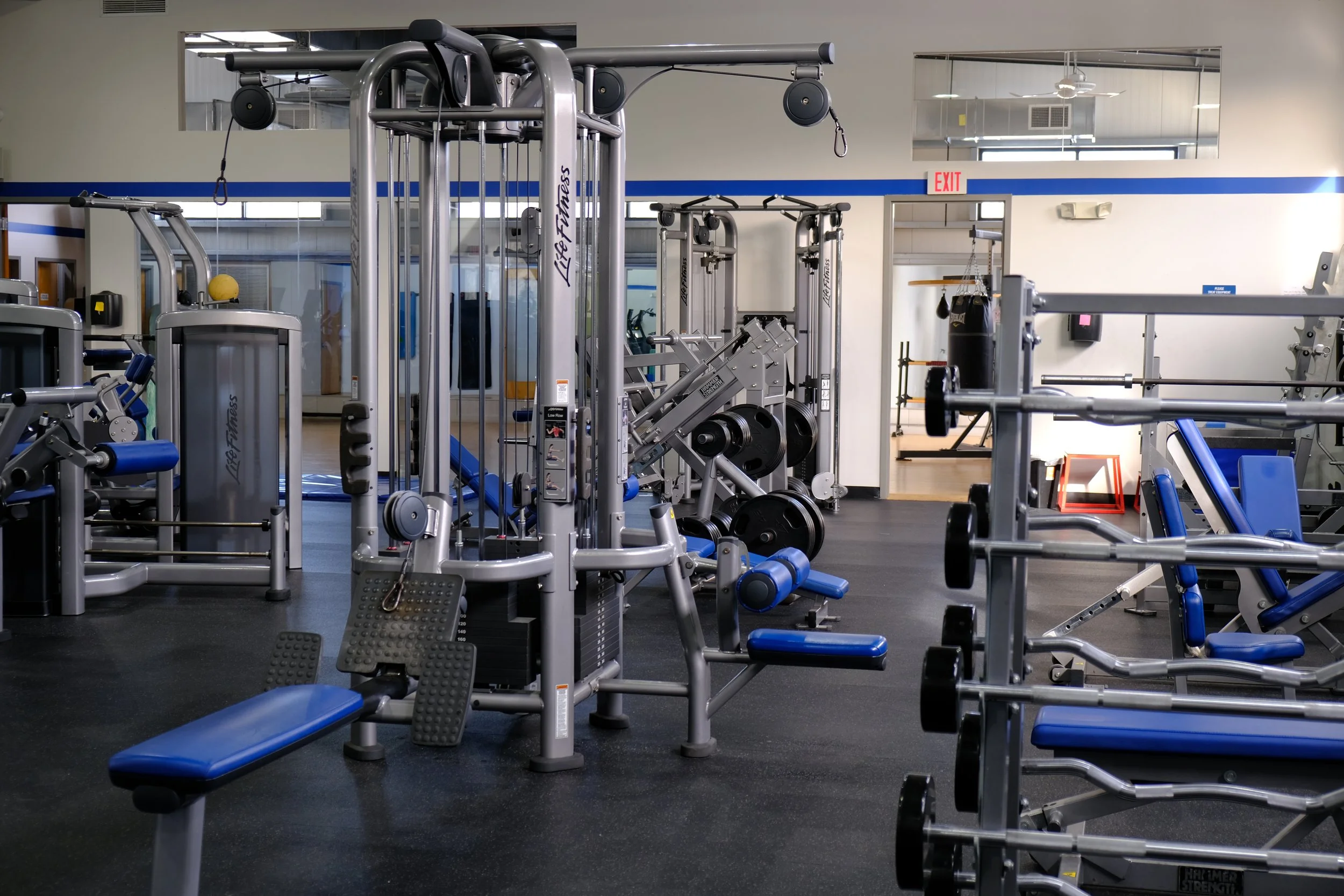 Empty gym with weightlifting and exercise equipment, including a bench, dumbbells, and resistance machines in a well-lit fitness center.