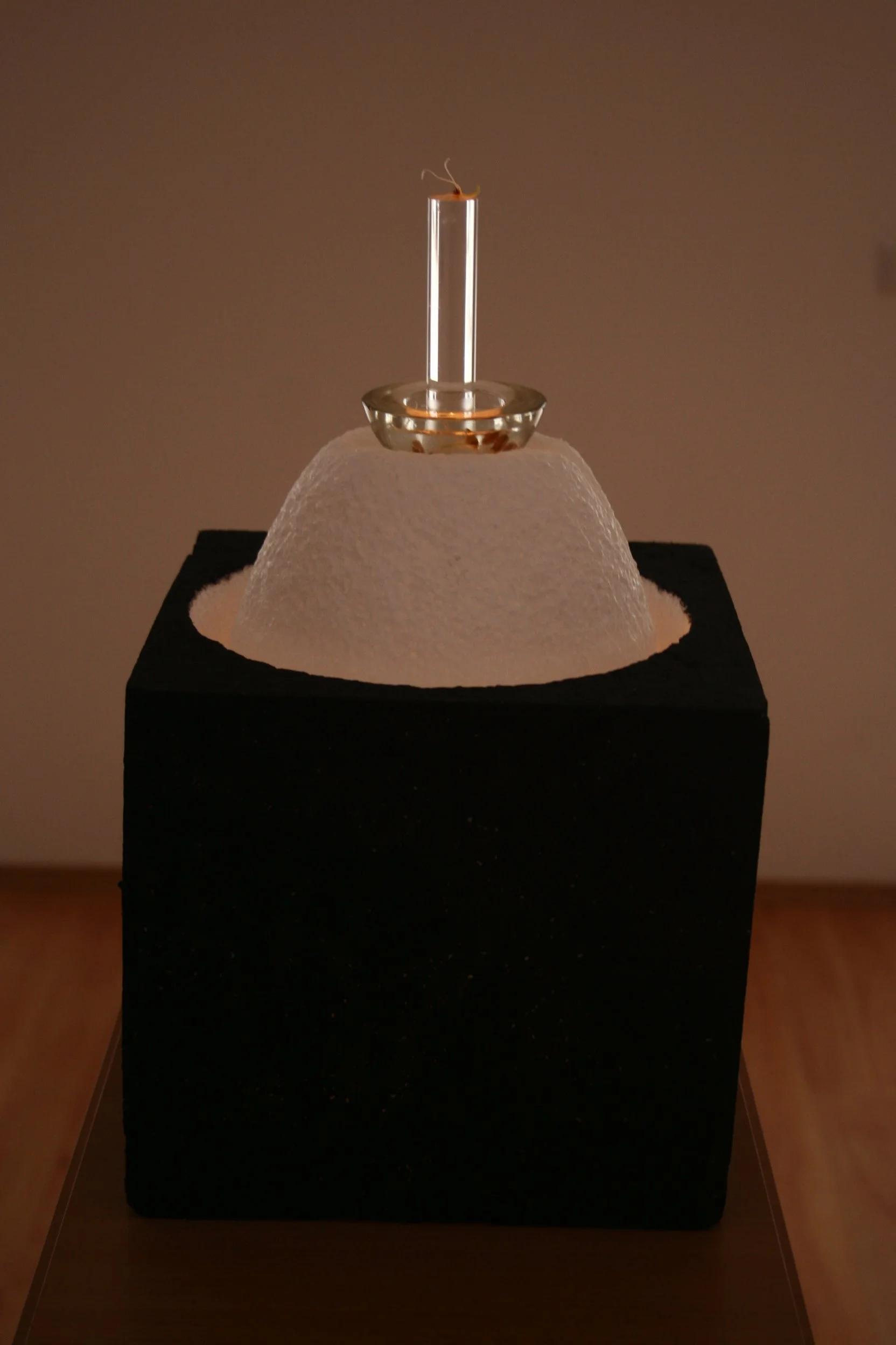 Minimalist sculpture featuring a textured white dome with a black base, topped by a small glass dish holding a clear glass tube, with a small orange object and thin wire at the top.