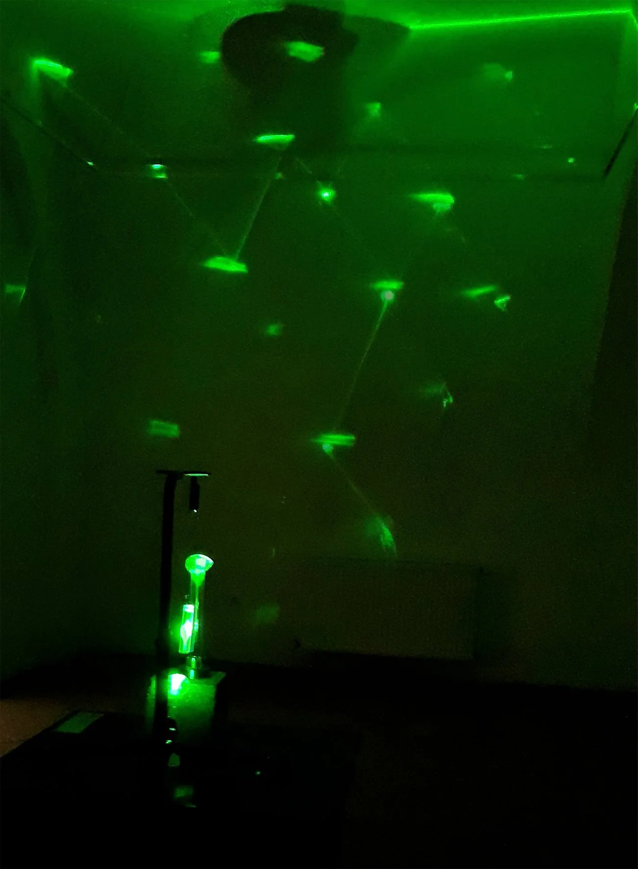 Green laser light projections on a wall and ceiling in a dark room, with a device emitting the laser light on a table.