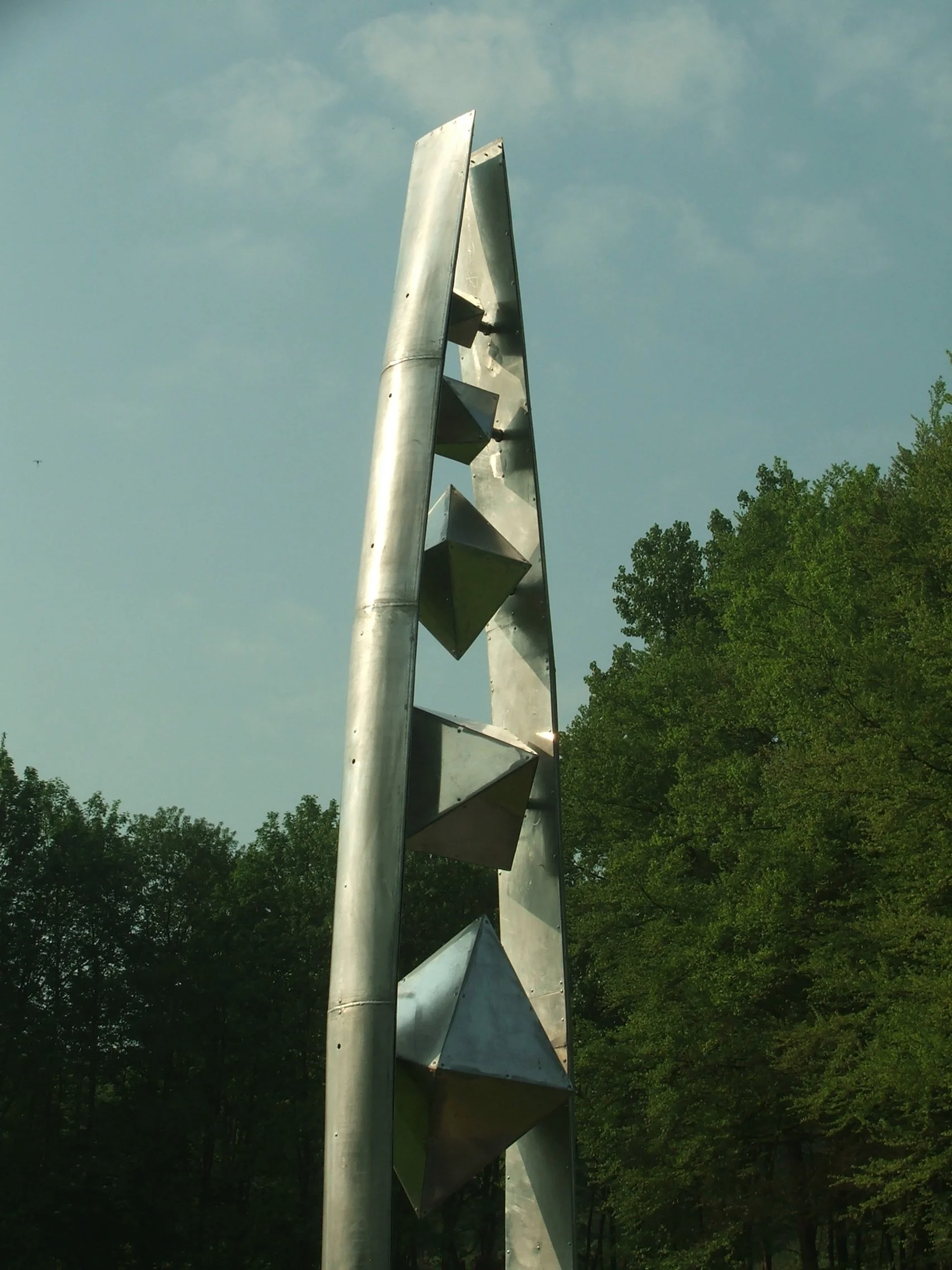 Tall metallic sculpture with geometric shapes, set outdoors against a background of trees and a cloudy sky.