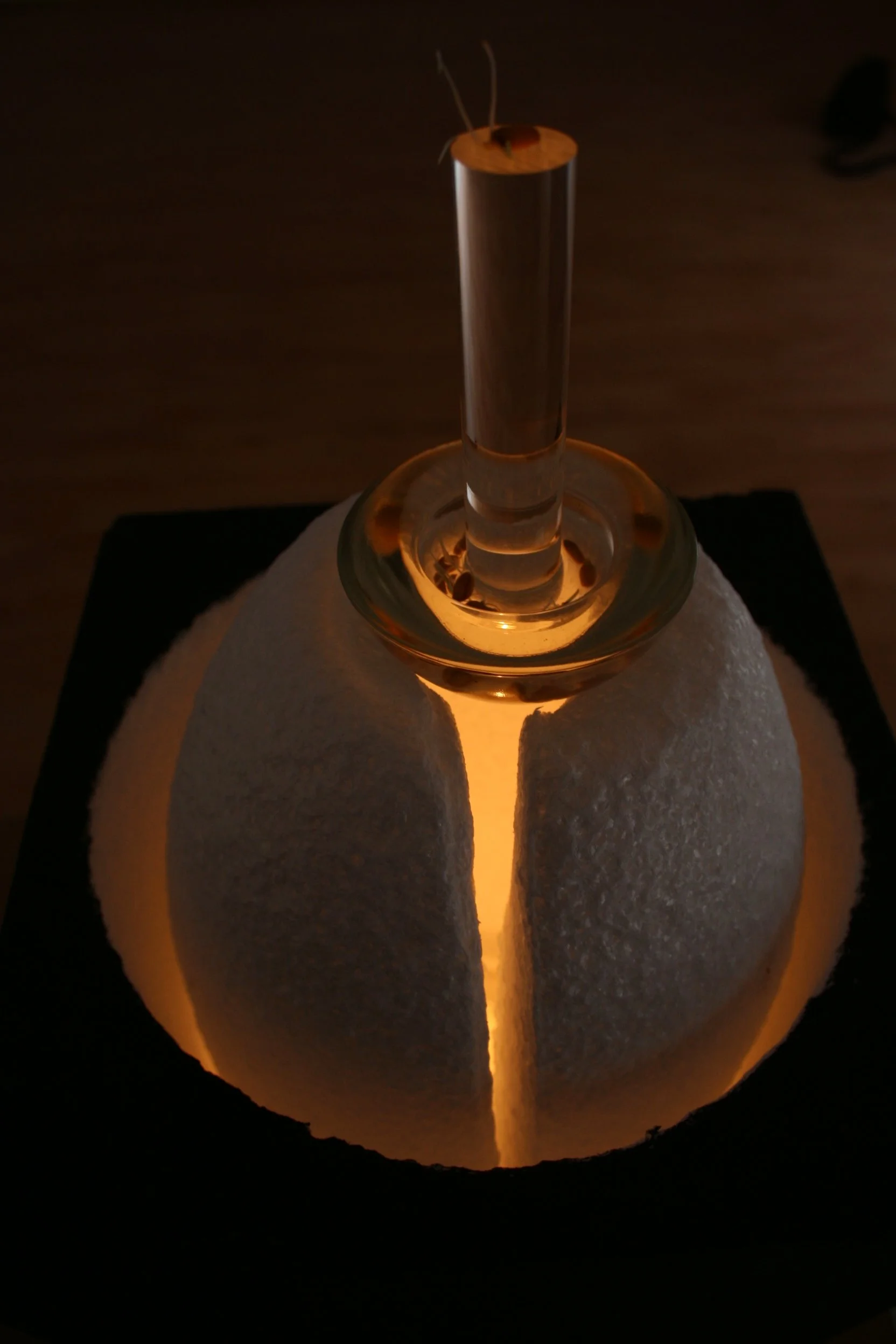 A white pumpkin with vertical cut open and a glass water pipe (bong) inserted into it, glowing from light within, on a dark surface.