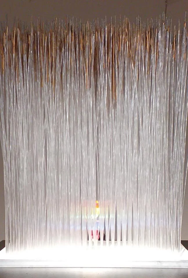 An abstract art installation featuring numerous long, thin, vertical metallic or plastic rods hanging from a top frame, creating a shimmering, curtain-like effect with a rainbow reflection at the bottom.