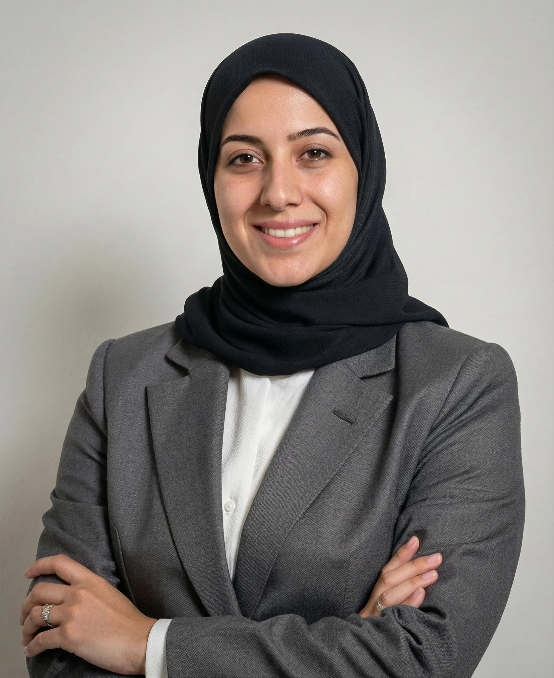 A woman wearing a black hijab, gray blazer, and white shirt smiling and standing with her arms crossed against a plain white wall.
