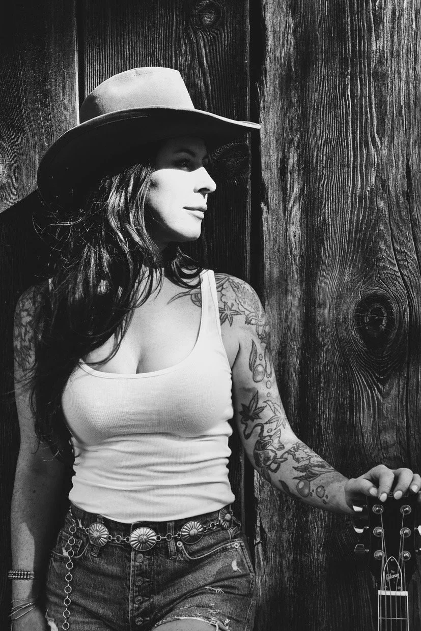 Black and white photo of a woman wearing a hat, tank top, and shorts, standing against a wooden wall, holding a guitar.