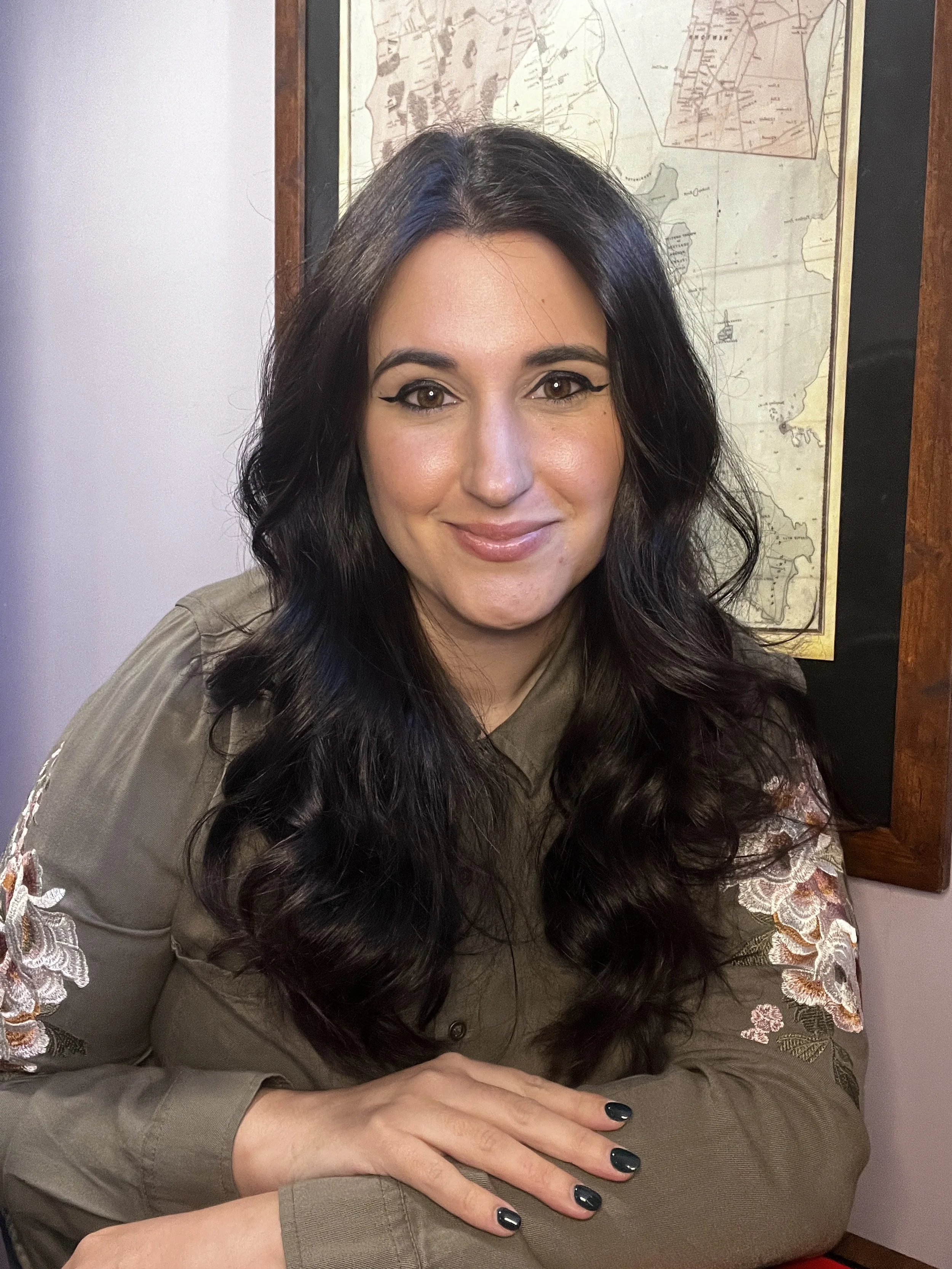 A woman with long dark hair and makeup sitting at a table, smiling, with a vintage map on the wall behind her.