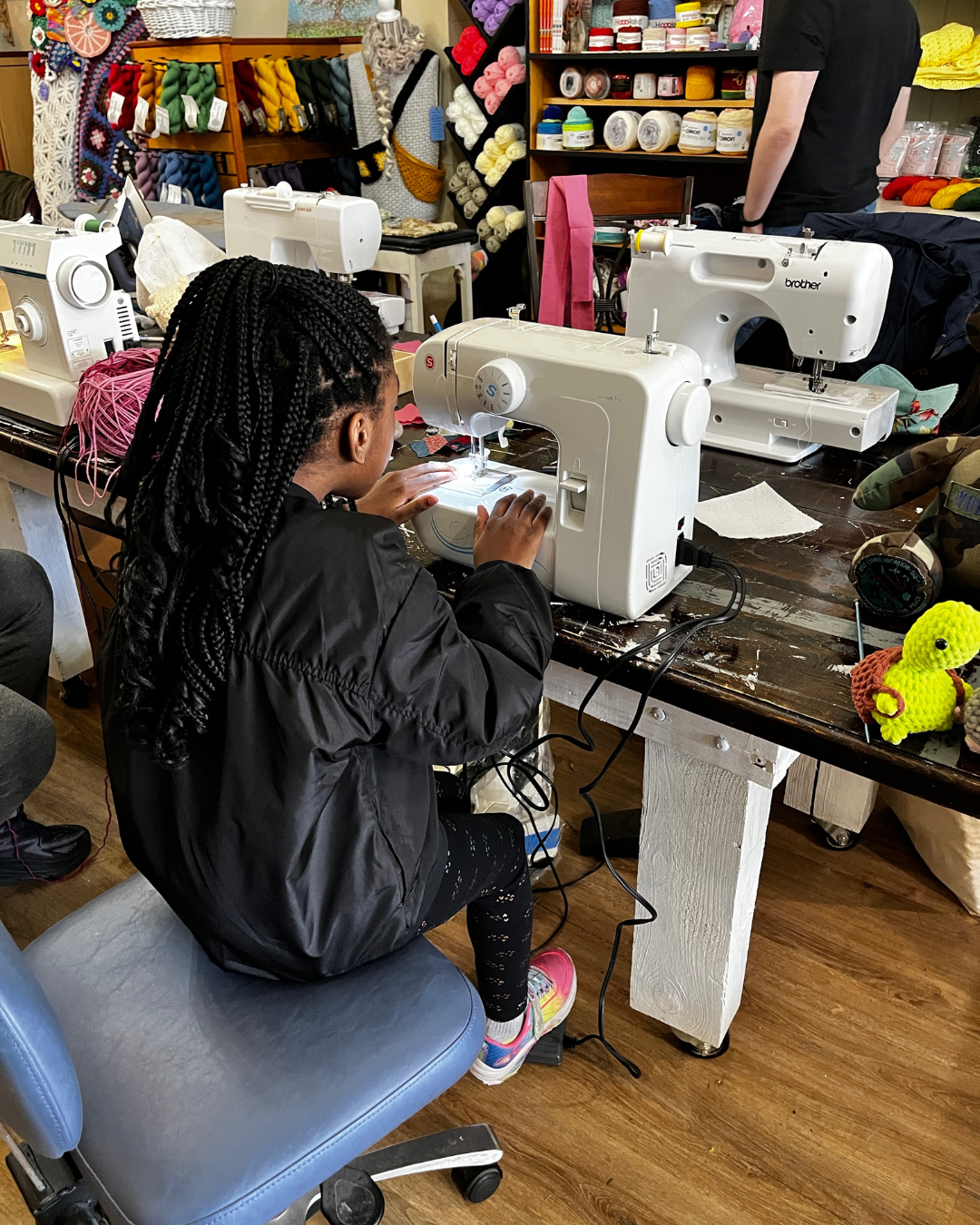 A girl sewing on a white sewing machine at a cluttered work table. Yarn, fabric, and craft supplies are visible around her.