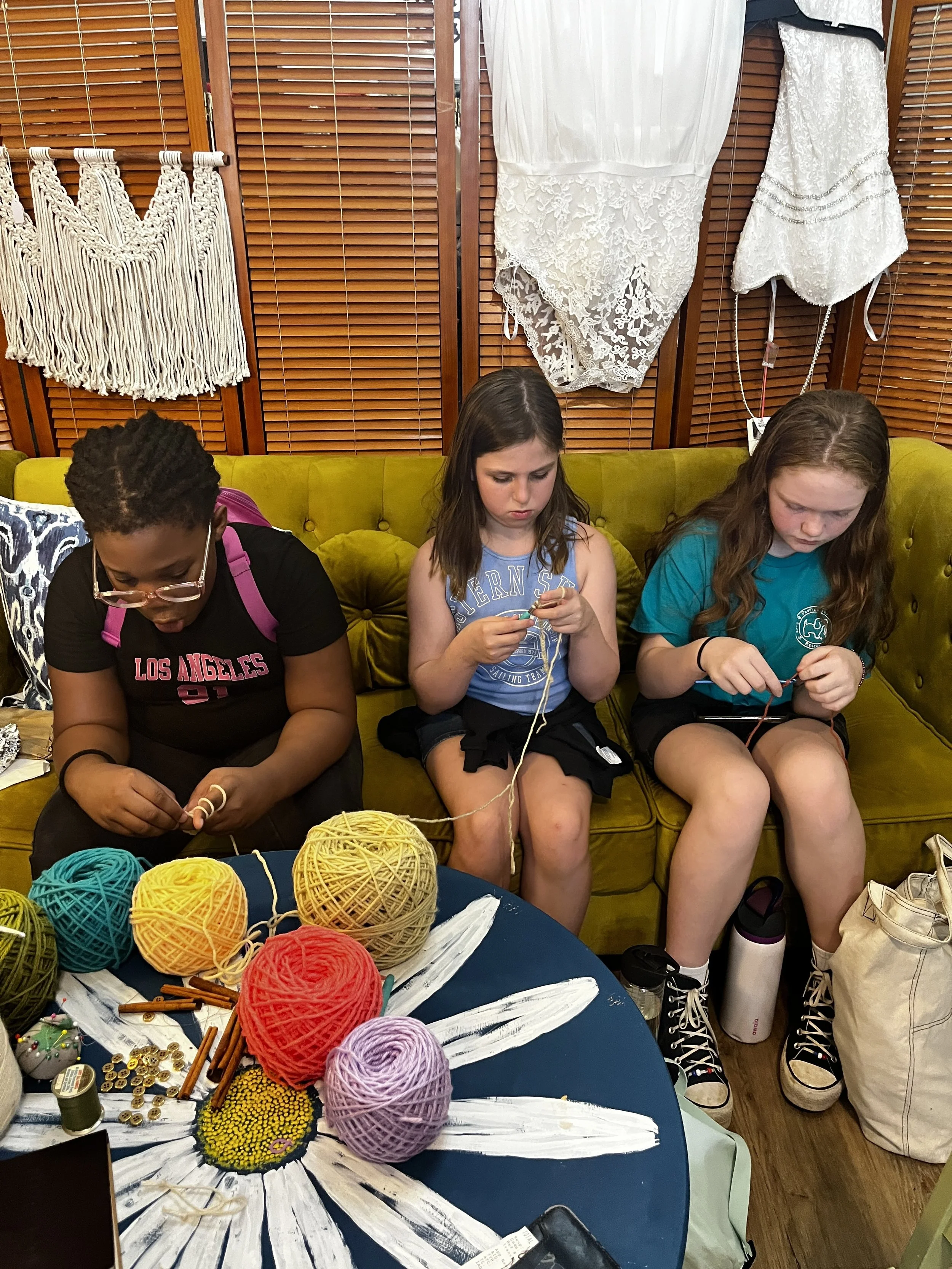 Three young girls sitting on a yellow sofa, engrossed in knitting or craft projects, with colorful yarn balls and craft supplies on a table in front of them, in a room with wooden shutters and white lace dresses hanging behind.