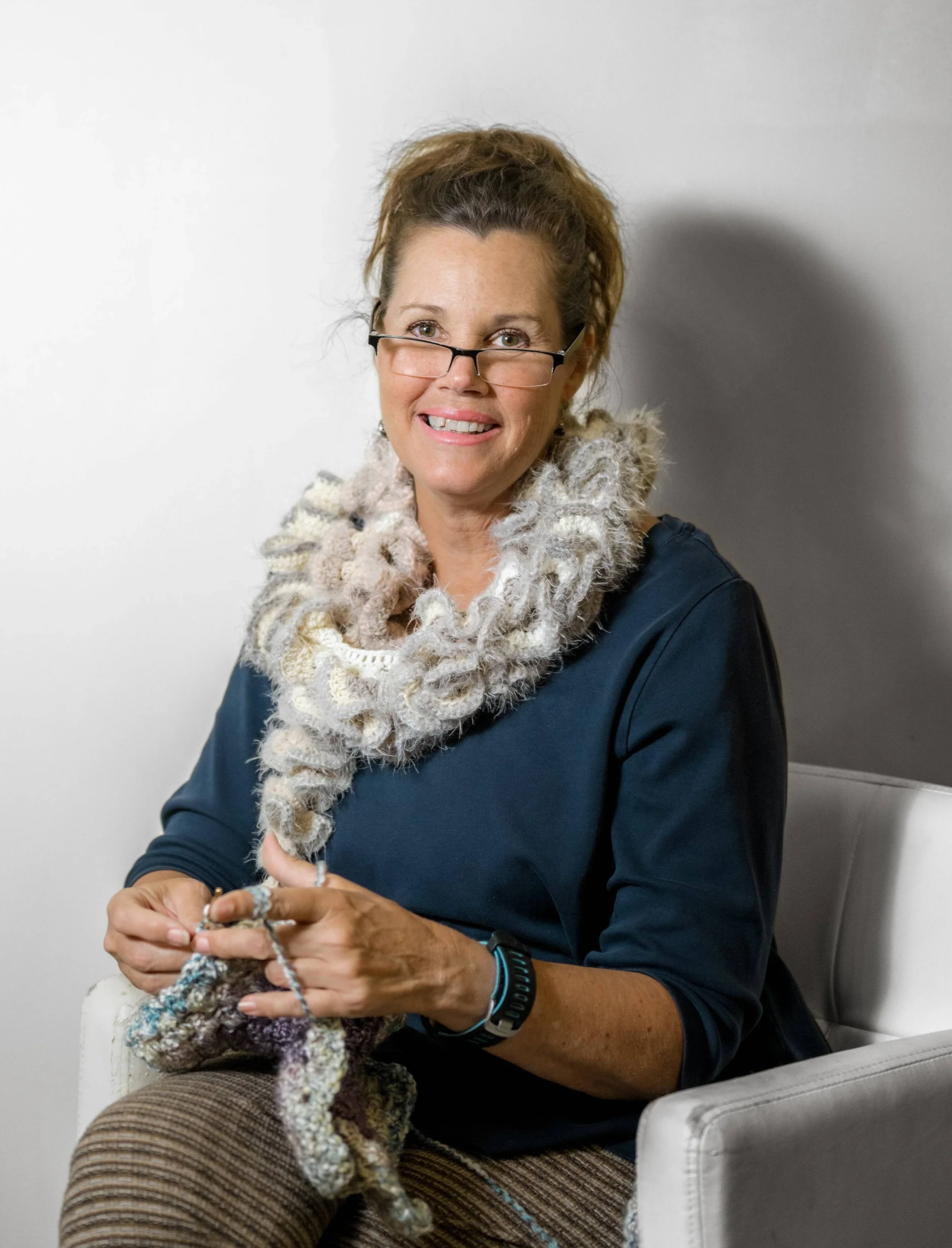 Woman knitting with a chunky scarf, wearing glasses, sitting in a chair, smiling, against a neutral background.