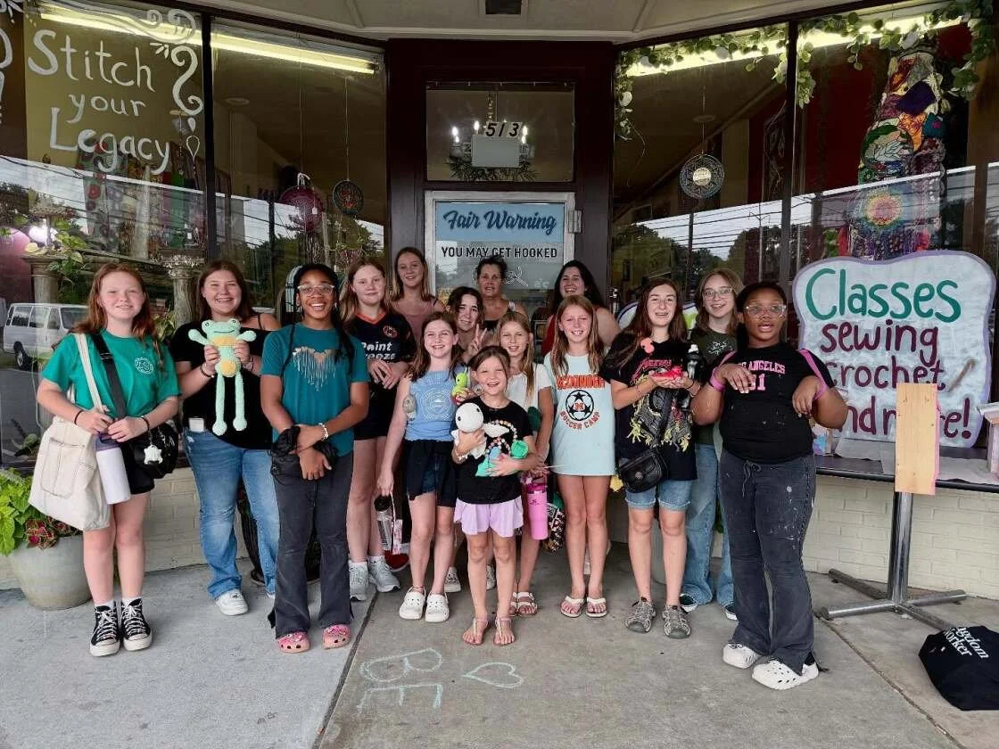 Group of young girls and women standing outside a storefront with signs promoting classes like sewing and crochet.