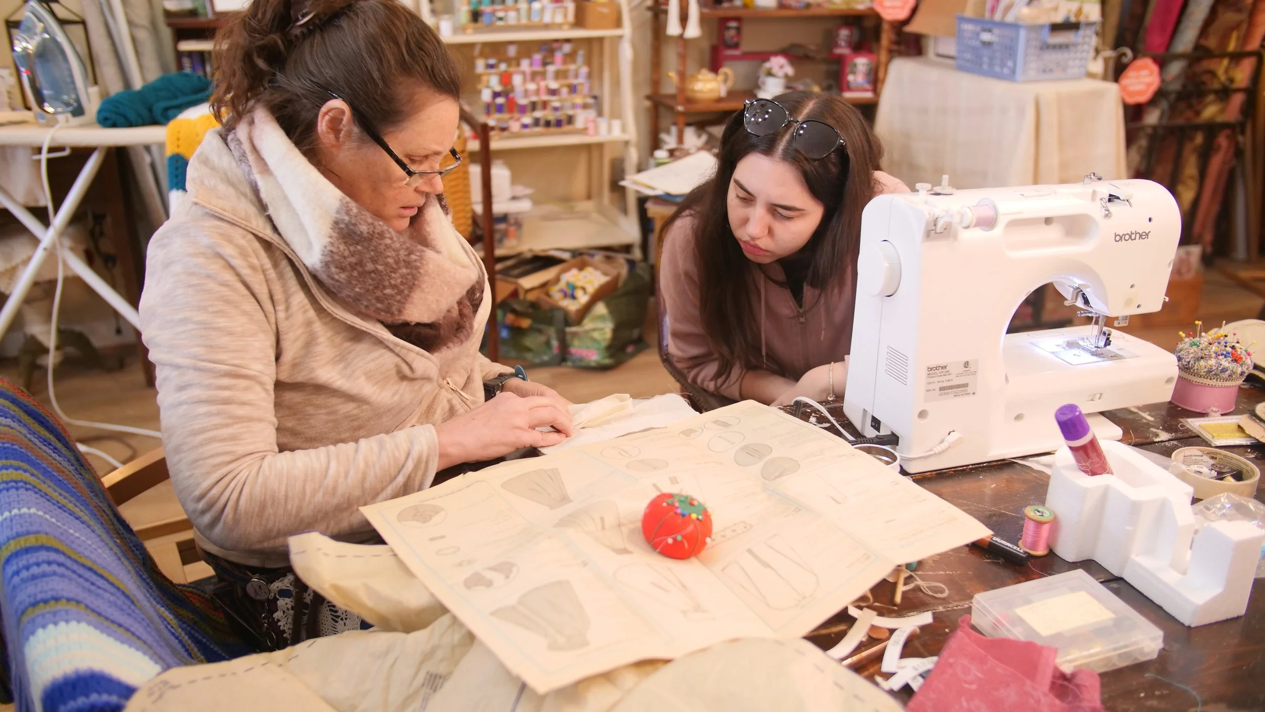 Two women working together on a sewing project in a craft room, with sewing materials and a sewing machine on the table.