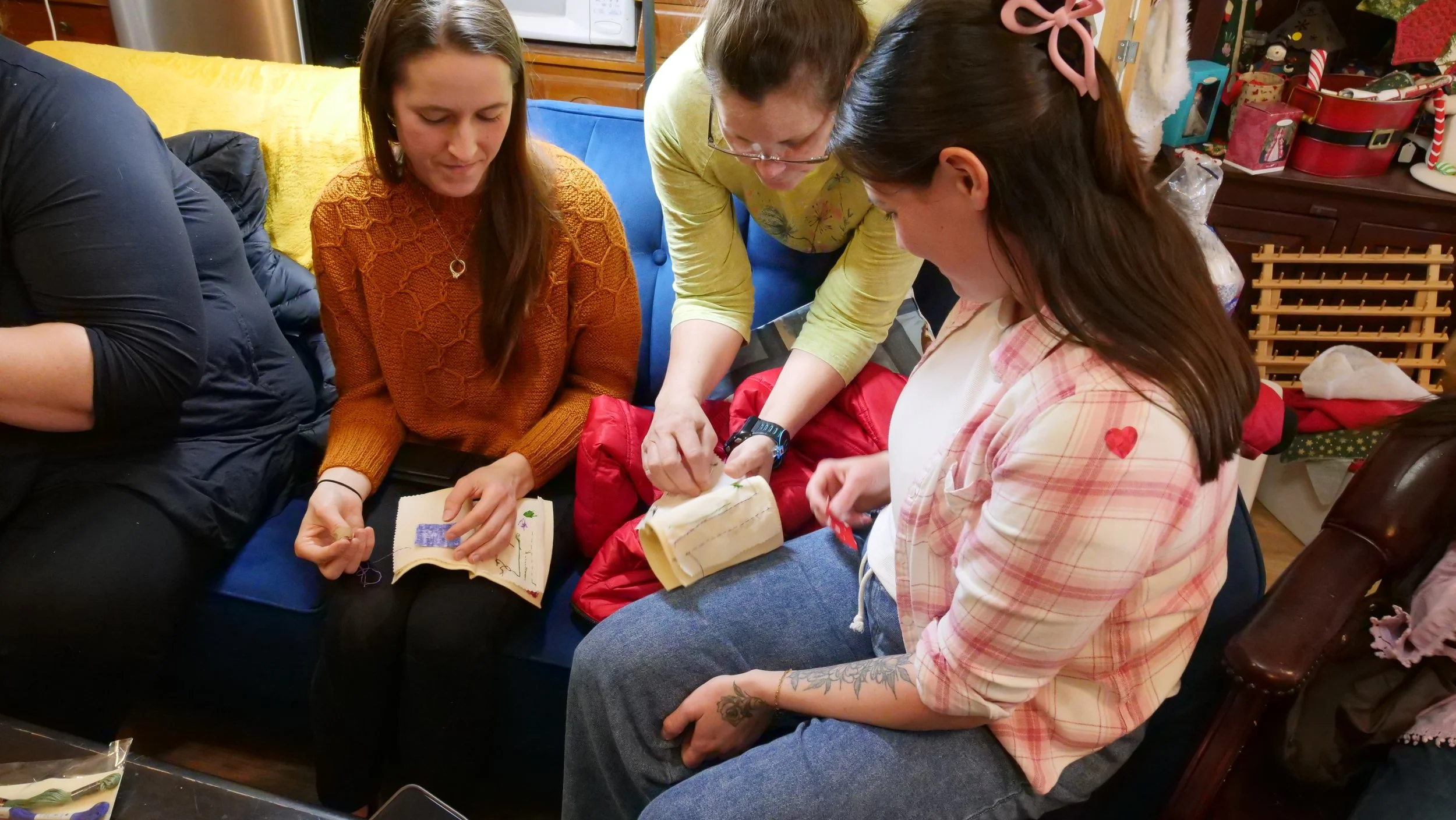 Group of women gathered around, working on embroidery and cross-stitch projects in a cozy, decorated room.