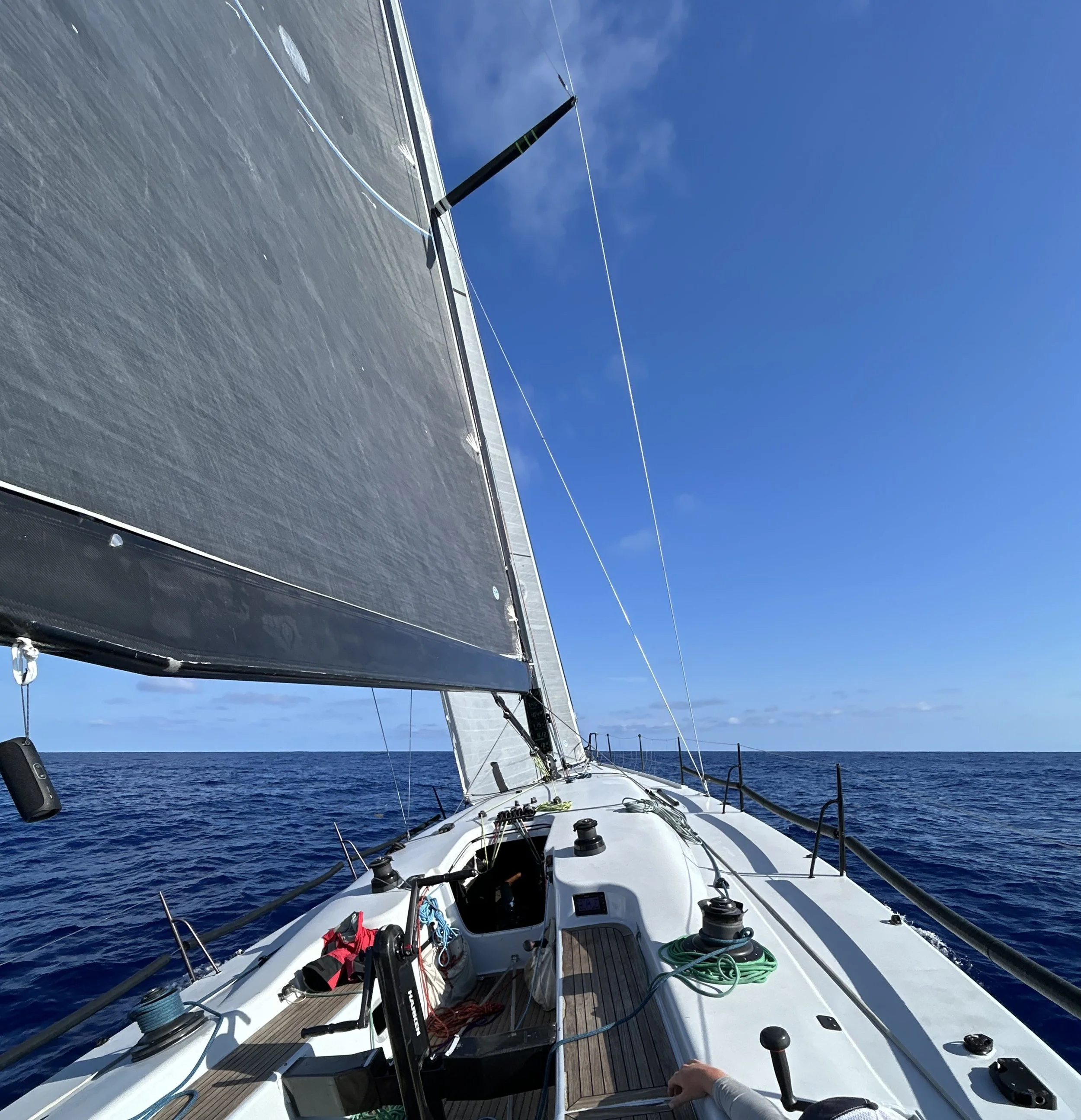View from a sailboat on open water showing the deck with fishing and sailing equipment, the sail mast, and the ocean extending to the horizon under a clear blue sky.