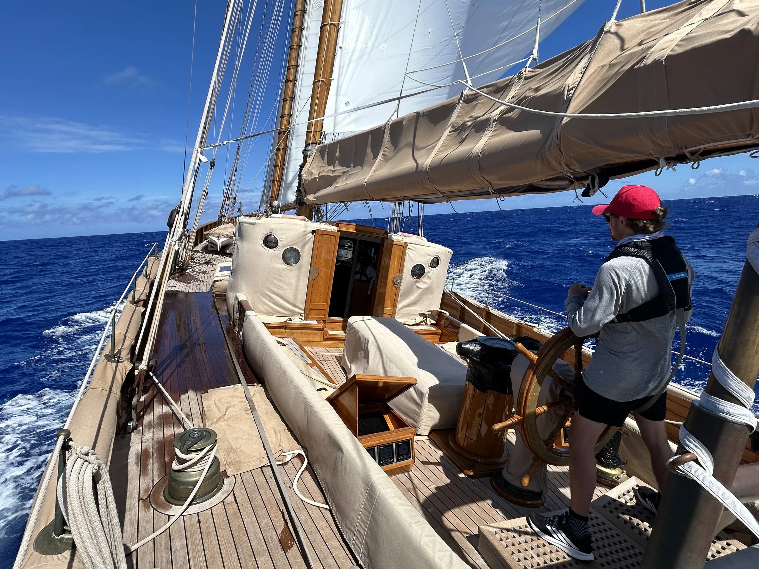 A person wearing a red cap and sunglasses steering a sailboat with a large beige sail, sailing on the blue ocean under a partly cloudy sky.